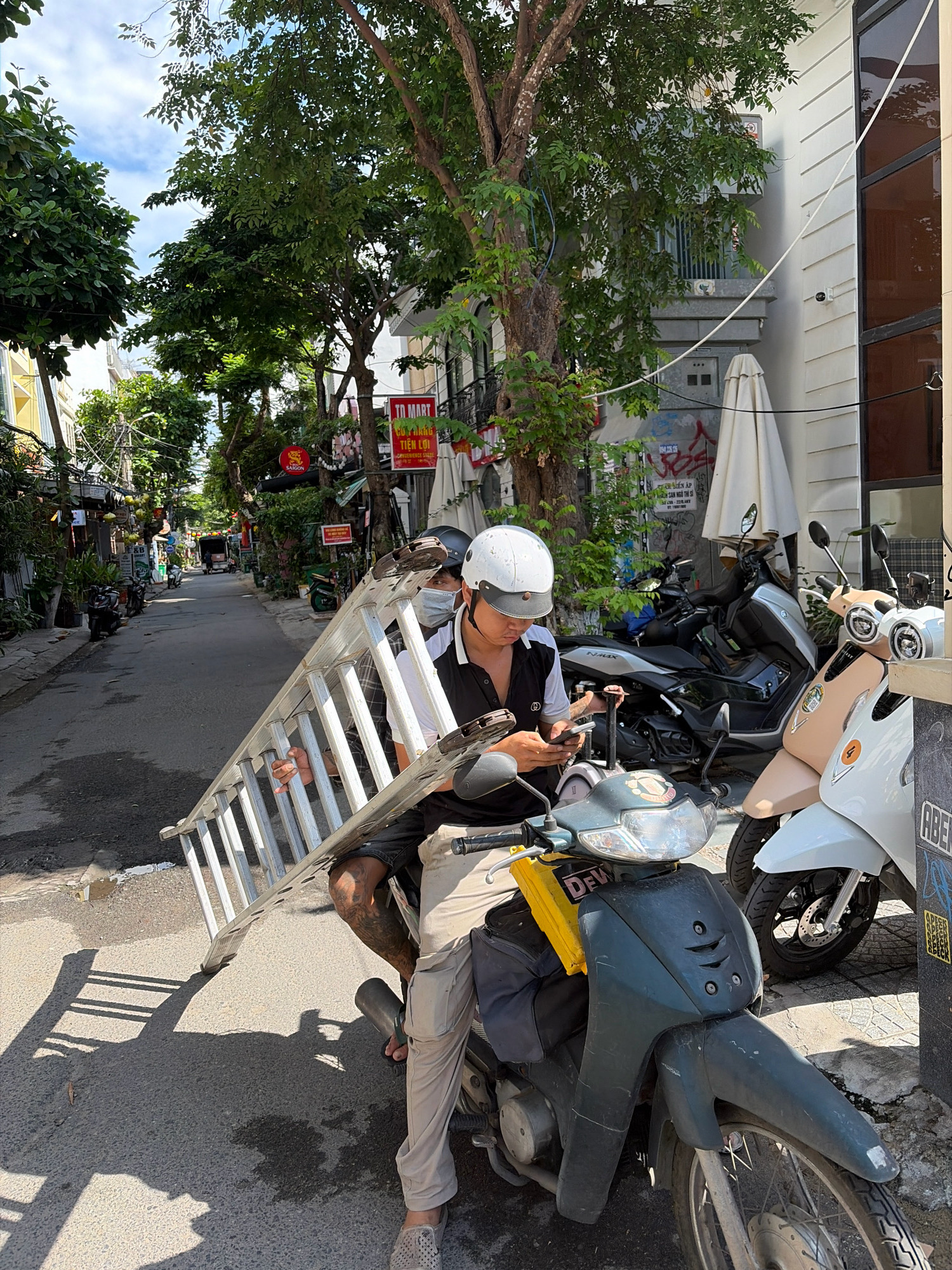 In a narrow street in Da Nang, Vietnam, a person wearing a white helmet and a face mask sits on a dark blue scooter. They are holding a long, silver ladder that extends diagonally across the frame, resting on the ground. The person is looking at their phone. Another person, also wearing a helmet and mask, is partially visible on the scooter, holding the ladder. Several other scooters and motorcycles are parked along the street, with buildings lining both sides. Green trees with lush foliage provide shade. The sky is partly cloudy.