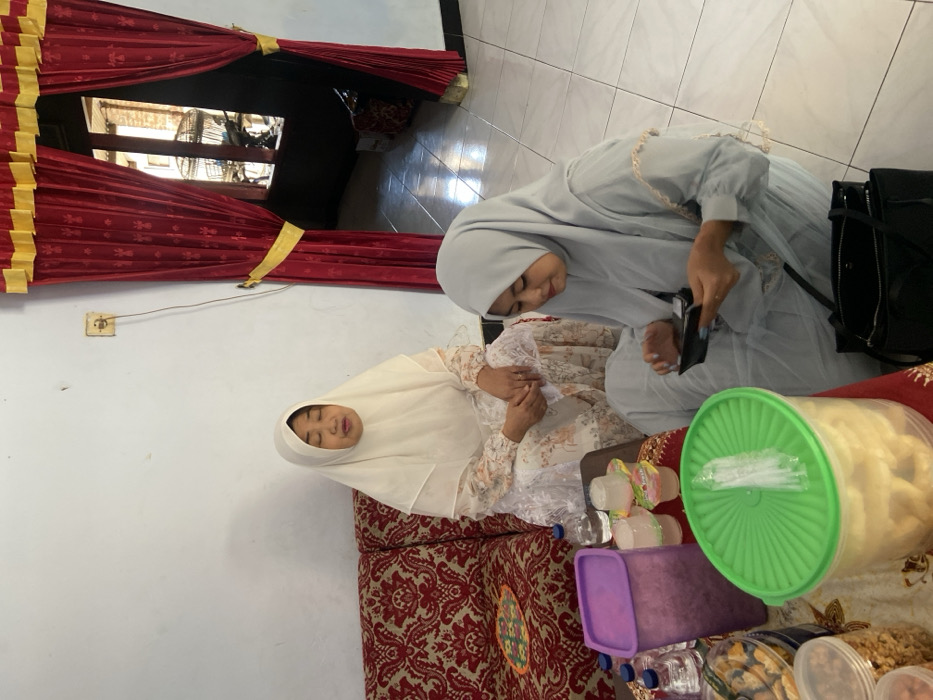 In an indoor setting in Tambakrejo, Indonesia, two women are present. On the left, a woman in a white hijab and a floral white dress is seated on a red patterned sofa, with her eyes closed or looking downwards, and her mouth slightly open. To her right, a second woman, wearing a light grey hijab and a matching light grey dress, sits cross-legged on the white tiled floor, looking down at a small black object, possibly a smartphone or wallet, which she holds with both hands. A small low table is positioned in front of the sofa, displaying several items: clear plastic bottles of water, several small clear plastic cups containing a translucent substance, a purple lidded container, and a large clear container with a green ribbed lid holding light-colored snacks. Other clear jars with snacks are also visible. A black handbag rests on the floor next to the woman in grey. The room features a plain white wall with a beige electrical outlet and an attached white cable. To the right, a doorway or window opening is framed by heavy red curtains with gold trim, tied back with gold sashes. Through this opening, an outdoor area with a birdcage is partially visible, indicating natural light entering the space.