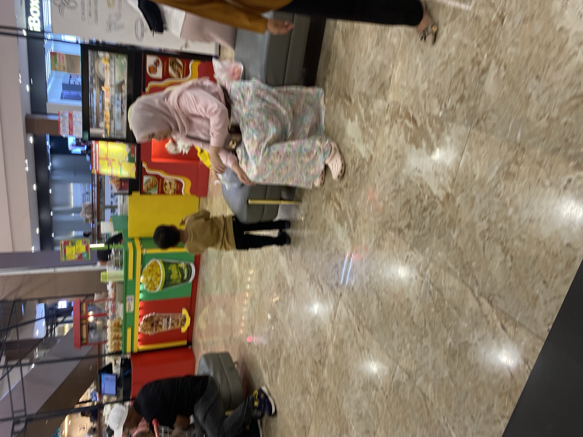 In a brightly lit indoor setting, possibly a shopping mall in Banda Aceh, Indonesia, a young child stands facing away from the camera, wearing a tan shirt and black pants. To the left, an adult wearing a white cap and black shirt is seated on a dark gray bench, with their legs crossed. To the right of the child, a woman wearing a light pink hijab and a floral patterned skirt is seated on the same bench, her hands resting in her lap. The floor is a polished marble tile with a light brown and beige pattern. In the background, a food stall with a red and green facade displays images of food items, and a glass display case with various food items is visible. The overall scene appears to be a casual moment in a public space.