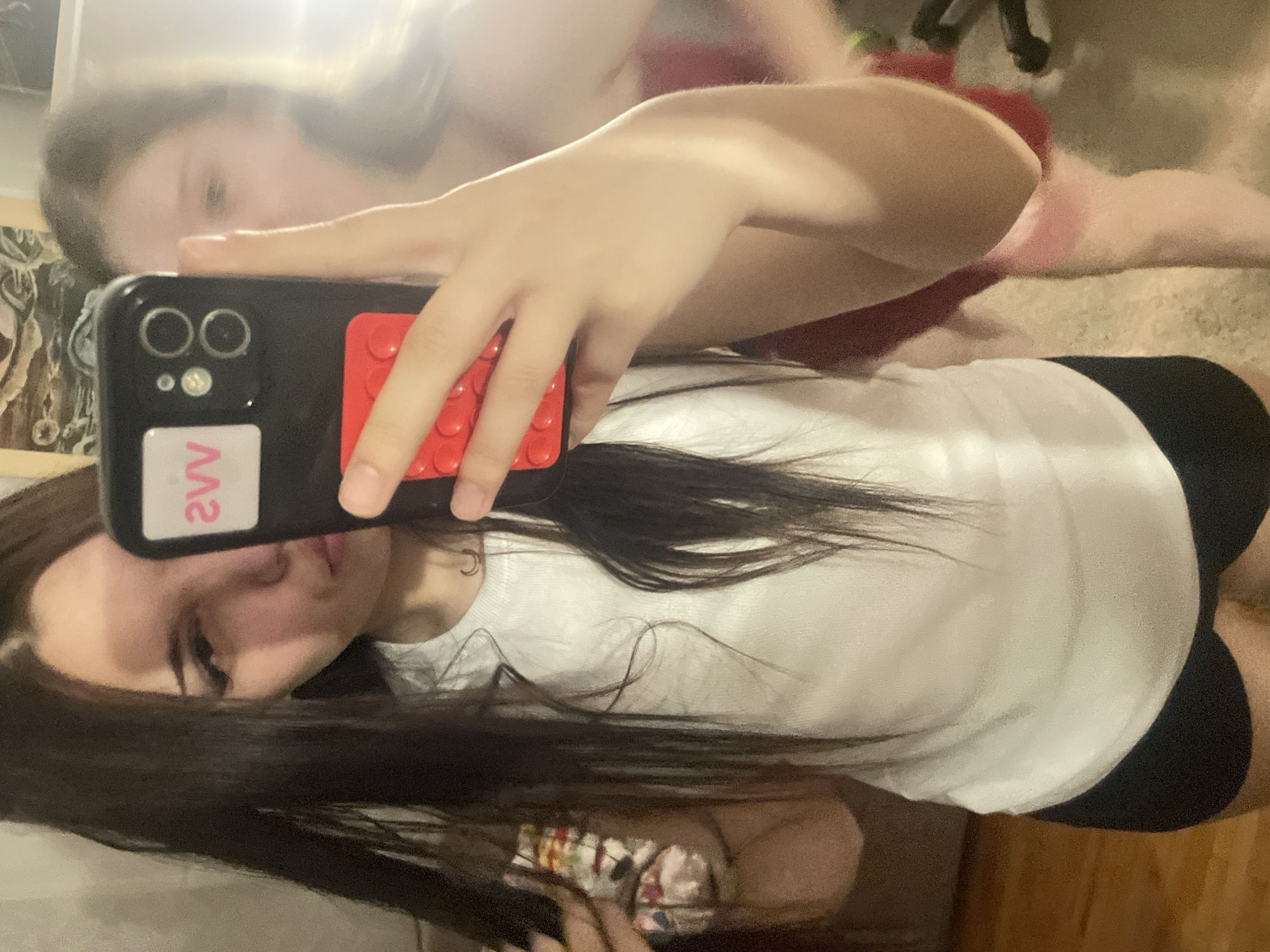 This mirror selfie captures two young women in an indoor setting, likely in Saint Petersburg, Russia. In the foreground, a young woman with long, dark hair is holding a black smartphone vertically to take the picture. She is wearing a white t-shirt and black shorts or leggings, with her face partially obscured by the phone. Her smartphone features a distinctive red, textured case that resembles Lego studs, and a rectangular sticker on the case displays the reversed text "SVV" in pink/red. The phone's dual cameras and flash are visible.
Reflected in the background is a second young woman with lighter hair, wearing a red top and possibly light-colored bottoms. She is lying in a relaxed pose on what appears to be a bed or floor, facing away from the direct view of the camera but clearly visible in the mirror's reflection. The overall scene conveys a casual and intimate moment between the two. The setting suggests a private room, possibly a bedroom, with glimpses of artwork on the left and a dark chair leg in the upper right background. The lighting is consistent with an indoor environment, possibly artificial, with a noticeable glare across the top portion of the mirror.