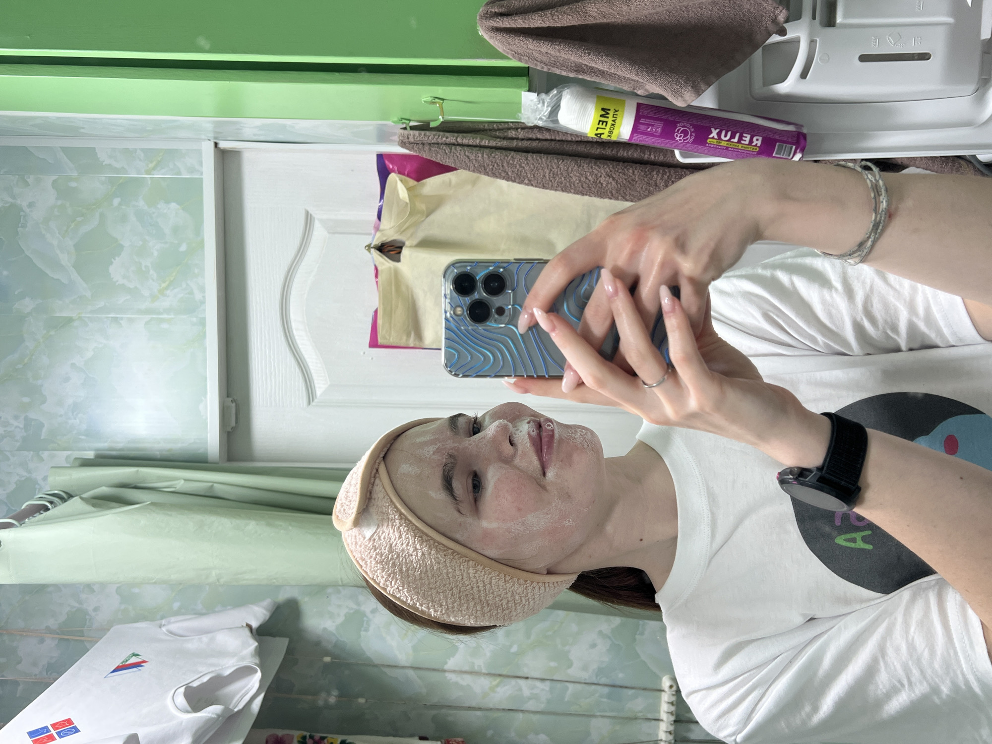 A woman is taking a selfie in a bathroom. The bathroom has light green marbled walls and a white door. She is wearing a white t-shirt with a graphic design, a light brown headband, and a smartwatch on her left wrist. She appears to be wearing a facial mask. In the upper part of the frame, there is a brown towel, a package of cotton pads ("BELUX"), and other toiletries. The woman's phone has a clear case with a blue wave pattern. The environment is domestic, and the photo was likely taken indoors. There is no concrete evidence to indicate it was taken in Chaikovsky, Russia.
