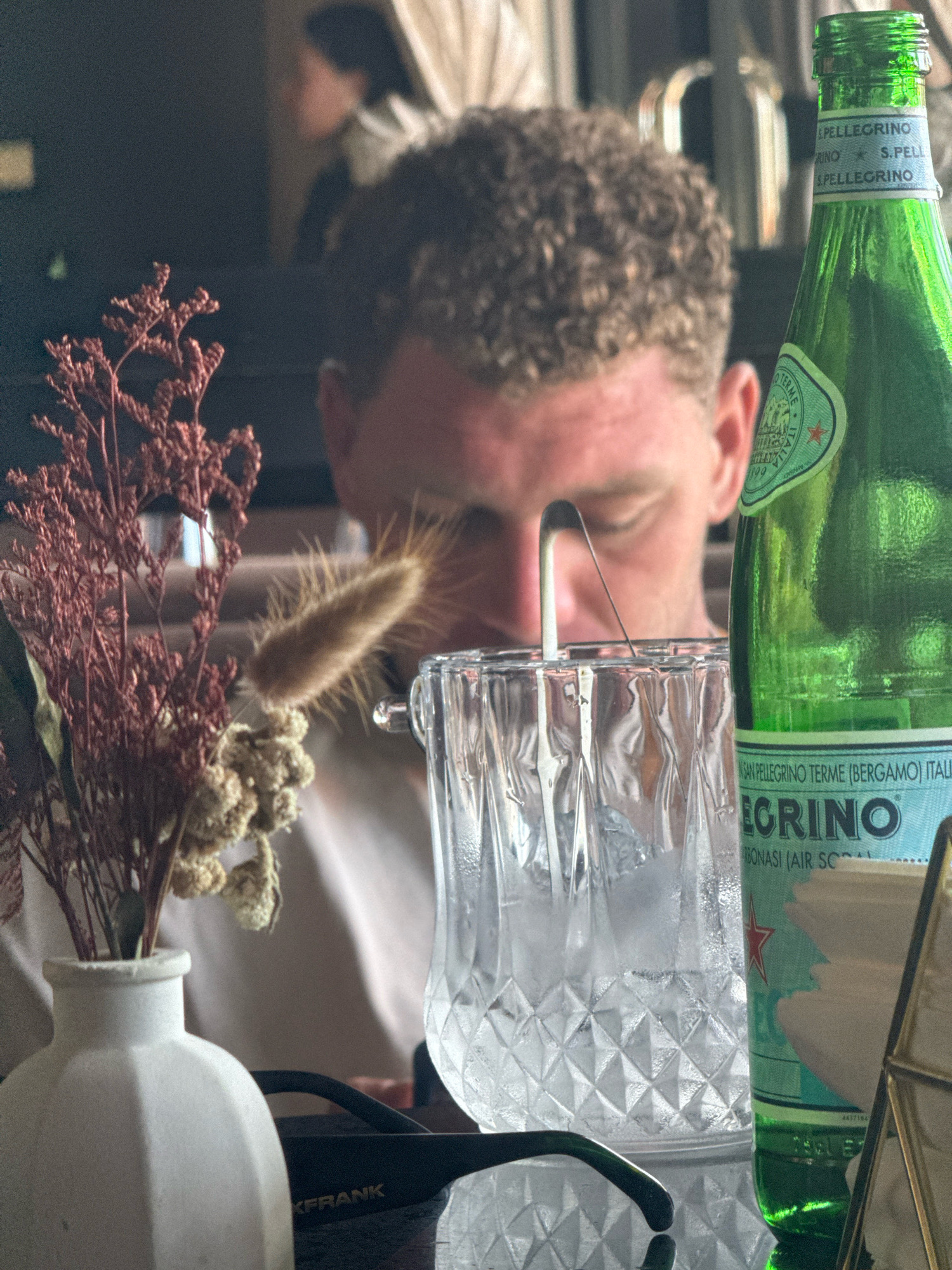 In the foreground of the image, a white ceramic vase holds a dried floral arrangement. To the right of the vase, a clear glass tumbler filled with ice and a straw is positioned. A green glass bottle of San Pellegrino water stands next to the tumbler. In the midground, a person with curly brown hair is looking down, their face partially obscured by the tumbler and straw. The background is out of focus and shows a dimly lit indoor setting with some beige curtains. The scene suggests a casual dining or social setting, possibly a cafe or restaurant. The image appears to be taken in Kuta Selatan, Indonesia.