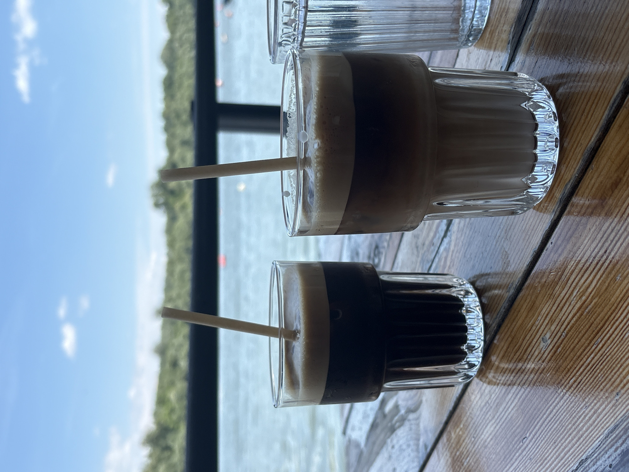The image shows a close-up of two glasses, likely filled with a layered beverage, on a wooden table. Each glass has a straw. Beyond the glasses, a window frames a view of water, vegetation, and the sky. The setting appears to be a cafe or restaurant in a waterfront location. Given the context of Zemun Urban Municipality, Serbia, it could be located near the Danube or Sava rivers in Zemun.