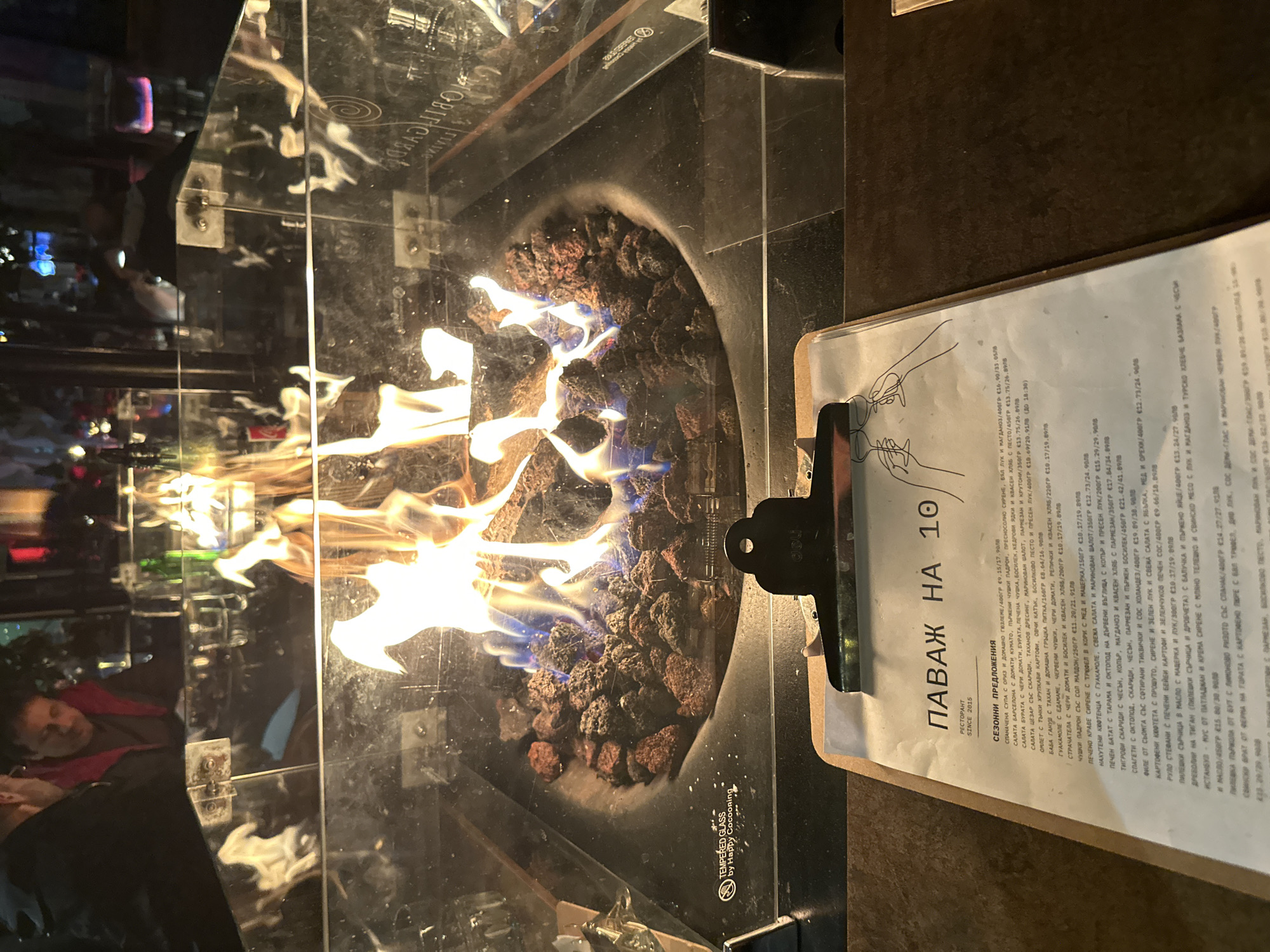 The image displays a nighttime scene inside or on the terrace of "Pavaj на 10" (Pavaj on 10), a restaurant located in Plovdiv, Bulgaria, as indicated by the Bulgarian menu title. The central focus is a square, black-framed gas fire pit with simulated logs and volcanic rocks, emitting bright orange and blue flames. The fire pit is enclosed by clear "TEMPERED GLASS by Happy Cocooning" panels. In the immediate foreground, a brown clipboard with a white paper menu is positioned on a dark tabletop. The menu, written in Bulgarian, is titled "ПАВАЖ НА 10" and notes "РЕСТОРАНТ SINCE 2015," featuring an illustration of two hands toasting wine glasses. In the blurred background, reflected in and visible through the glass, are two indistinct male figures seated opposite the camera; one wears a dark jacket, and the other a red/maroon jacket. The environment is dimly lit, with reflections of ambient lights and out-of-focus colored lights visible in the glass panels surrounding the fire.
