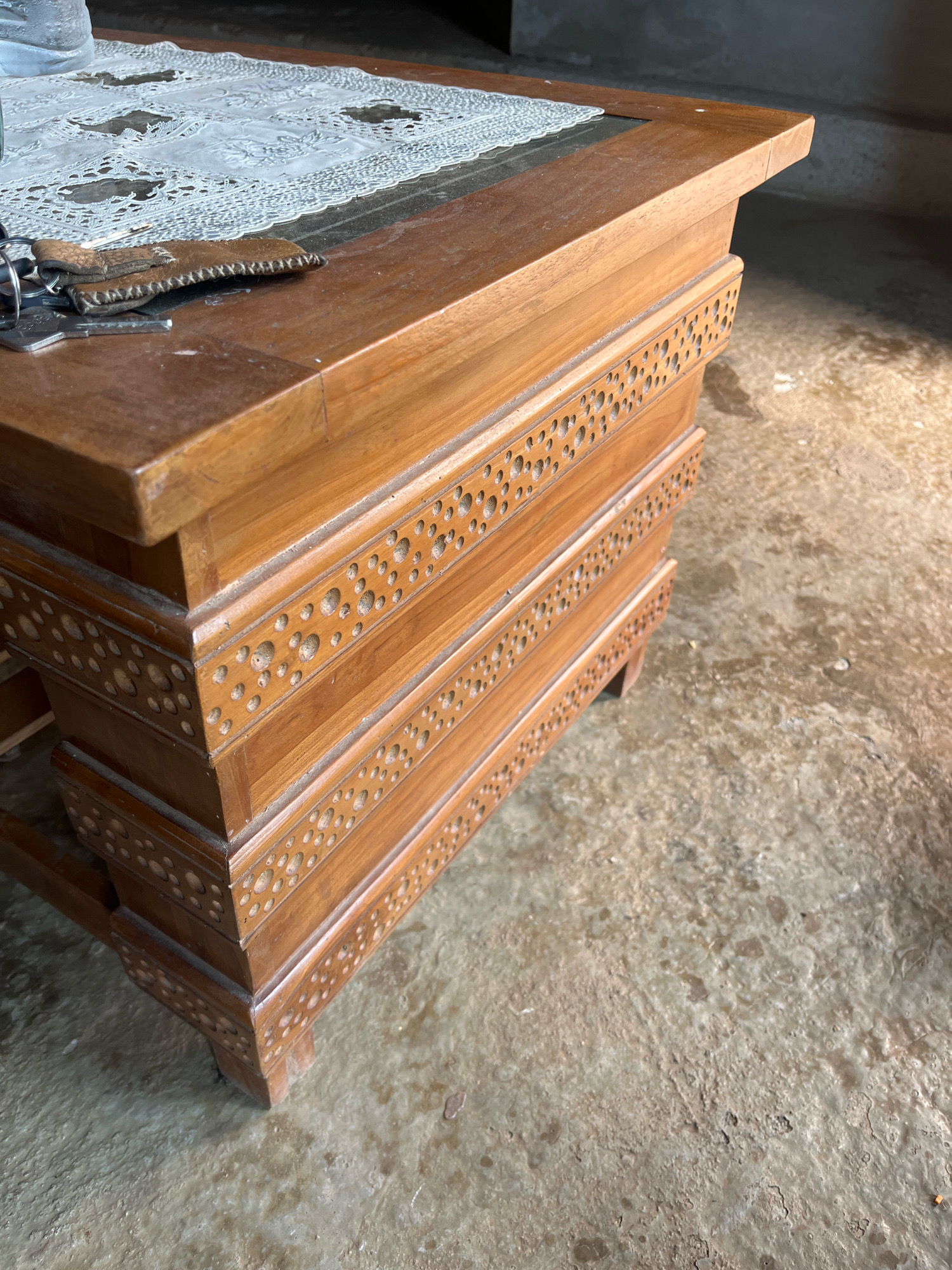 A wooden table with intricate carved patterns is situated on a concrete floor. A white lace doily covers a portion of the table's surface, and a set of keys with a brown leather fob rests on the table. The environment appears to be indoors, possibly a residential setting in Sinar Mulyo, Indonesia, with a wall visible in the background and sunlight casting shadows on the floor.