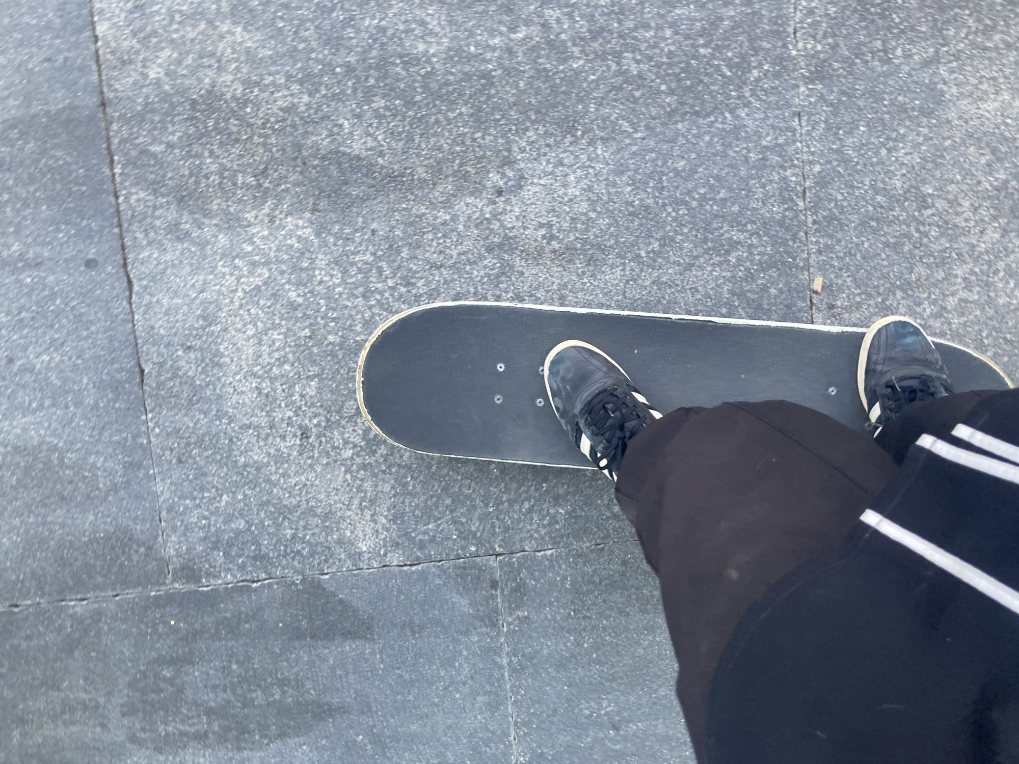 The image shows a person's legs and feet on a skateboard. The skateboard is black and positioned on a gray, textured pavement. The person is wearing dark pants with white stripes down the sides and dark sneakers with white accents. The viewpoint is from above, looking down at the skateboard and the person's lower body. The pavement appears to be made of large, square tiles with visible cracks and some areas of dampness, suggesting an outdoor urban environment. There are no other people or objects clearly visible in the frame. The overall impression is a candid shot of someone preparing to skate or in the process of skating.