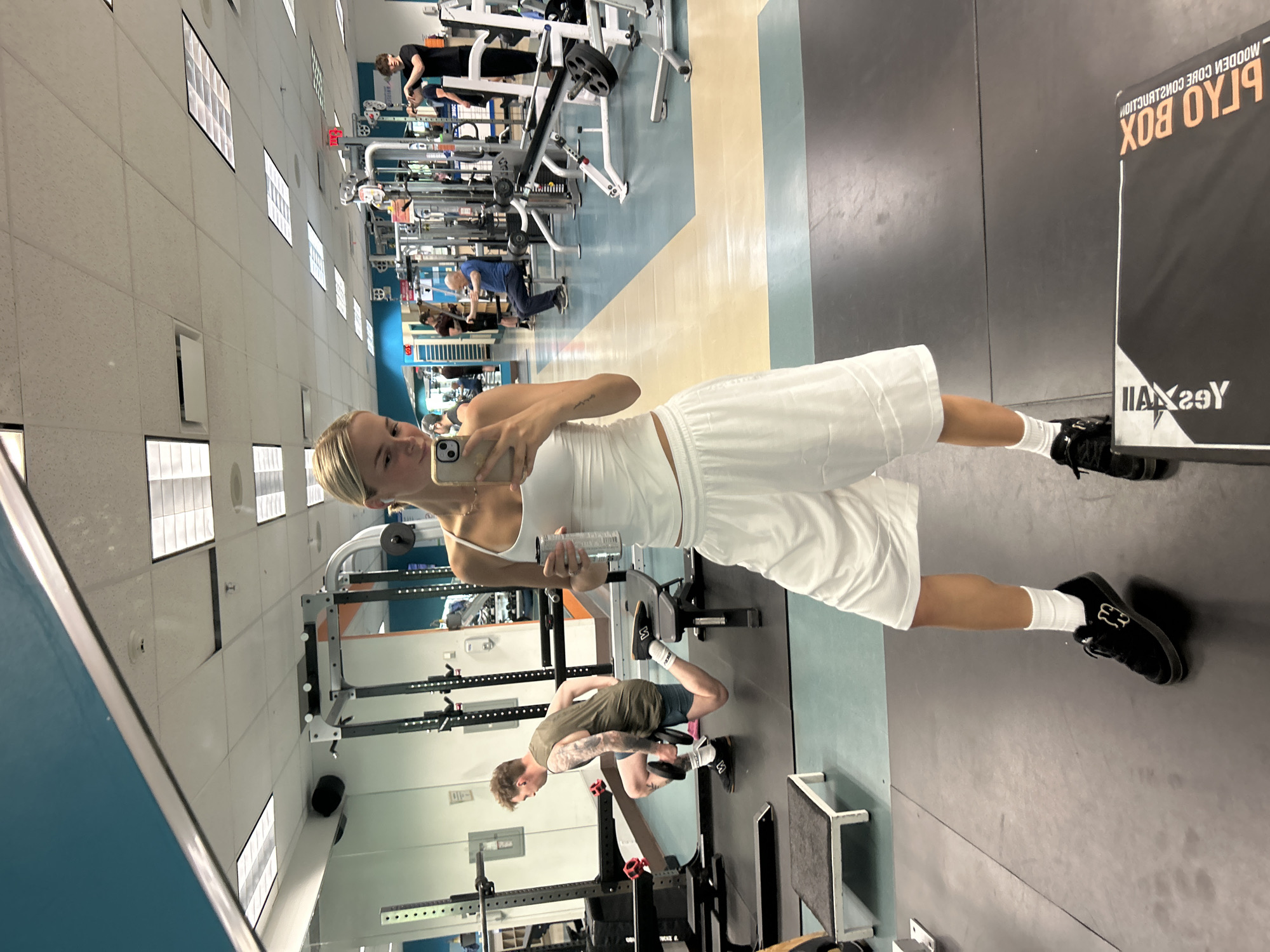 A young Caucasian woman with blonde hair tied back in a ponytail, wearing a white tank top, white athletic shorts, white socks, and black sneakers, stands in a gym, taking a selfie in a mirror. She holds a clear water bottle in her left hand and her phone in her right. In the background, through the mirror's reflection, several other individuals are visible engaged in various exercises. To the woman's left, a man wearing a sleeveless olive green shirt and blue shorts is performing a lunge with a weight. Further back, other gym equipment and patrons are present. The gym environment includes weight racks, machines, and a tiled floor. The woman is positioned centrally in the foreground of the image, facing the mirror. The scene is indoors, in what appears to be a public fitness facility. The location is not specified to be in Whistler, Canada.