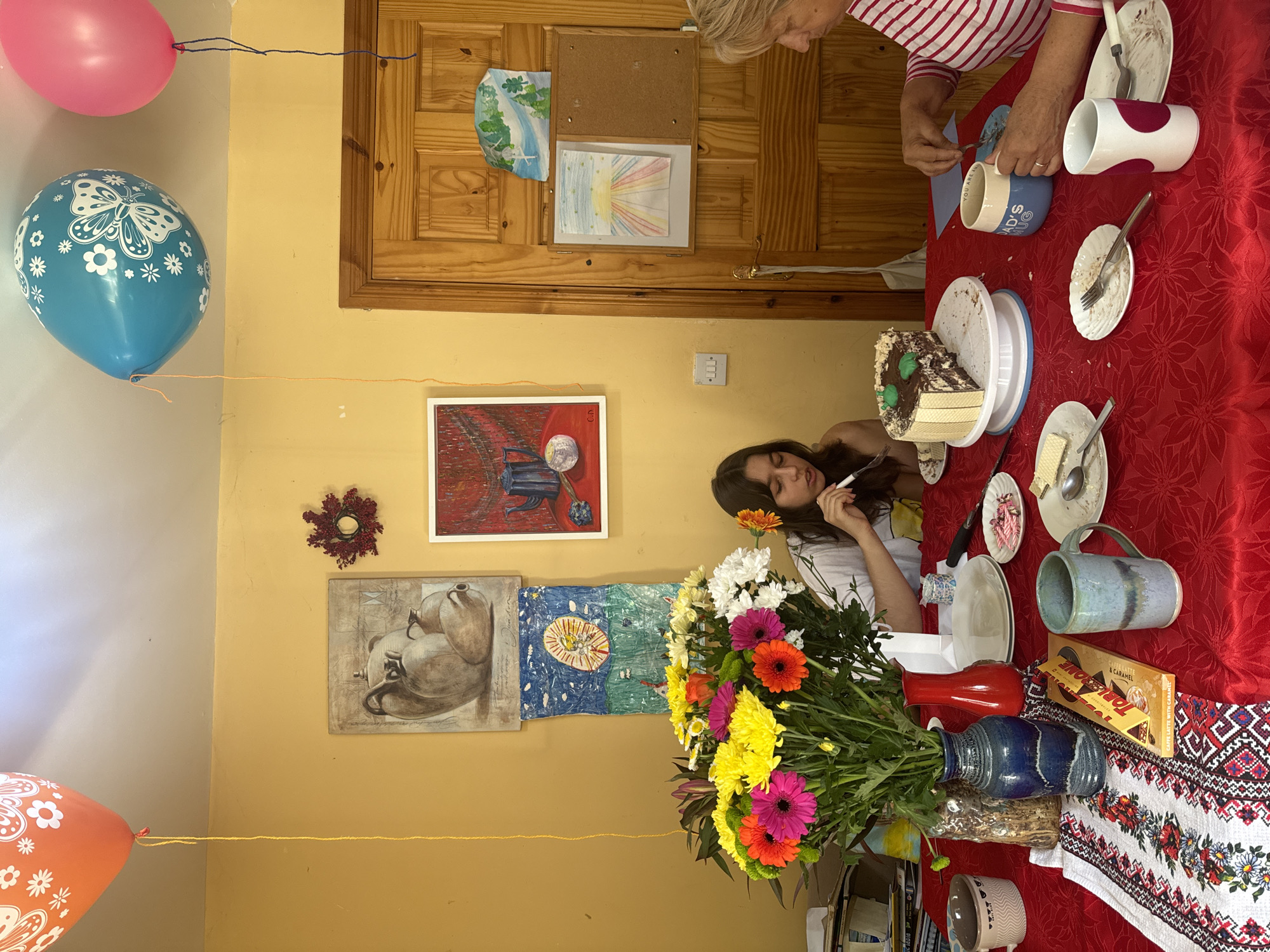 The image depicts an indoor scene, likely a dining room, set for a celebration. A red tablecloth covers the table, adorned with a floral-patterned runner. On the table, a partially eaten chocolate cake sits on a white cake stand. Several plates with cake remnants, forks, spoons, and some wrapped sweets are also present. A bouquet of colorful flowers in a blue vase and a red vase adds a festive touch. A young girl with dark hair, wearing a white shirt and an orange flower in her hair, is seated at the table, holding a fork and appearing to eat. Beside her, an older woman with blonde hair, wearing a striped pink and white shirt, is also seated, holding a fork and engaged with a mug that reads "DAD'S MUG." Several other mugs and cups are scattered around the table. In the background, yellow walls are decorated with framed artwork and a wreath. Three balloons, one orange, one blue, and one pink, are tied to the ceiling. A wooden door is visible on the right side of the frame, with various drawings and a bulletin board attached to it. There are no identifiable landmarks or specific indicators to place this scene within the city of Waterford, Ireland.