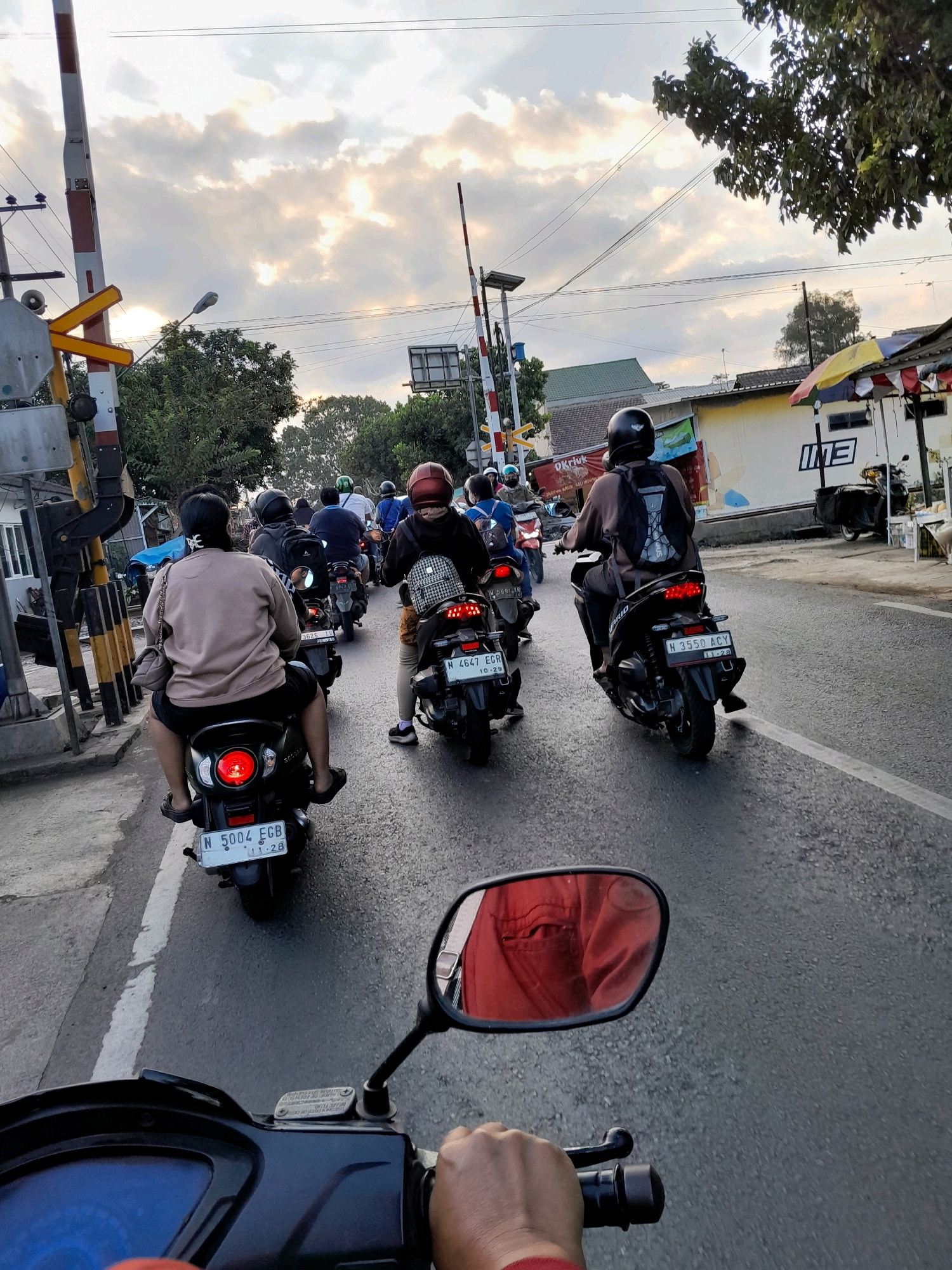 The image captures a traffic scene in Pakisaji, Indonesia, likely near a railway crossing, indicated by the presence of a red and white railway barrier in the foreground. A line of motorcyclists is queued on an asphalt road, appearing to be waiting. The sky is overcast with dramatic clouds, suggesting it could be late afternoon or early evening.
In the foreground, the perspective is from the rider of the first motorcycle, with a partial view of the handlebars and the rider's hand. Directly in front, another motorcyclist is visible, wearing a light brown long-sleeved shirt and dark shorts, with a black bag slung over their shoulder. This rider is looking ahead, and their motorcycle has a visible license plate "N 5004 FGB".
Behind them, several other motorcycles and riders are in the queue. The second rider from the front is wearing a red helmet and a backpack. The third visible rider has a black helmet and is wearing dark clothing, with a backpack. Further back, more motorcyclists are present, some with helmets, others without.
To the right of the road, there are buildings with some commercial signage, including what appears to be "INE" in large letters on one building. A small stall with a colorful umbrella is also visible. Trees line the sides of the road, and power lines stretch across the sky. The overall impression is a typical urban traffic situation in Indonesia, with a focus on the motorcycles and their riders.