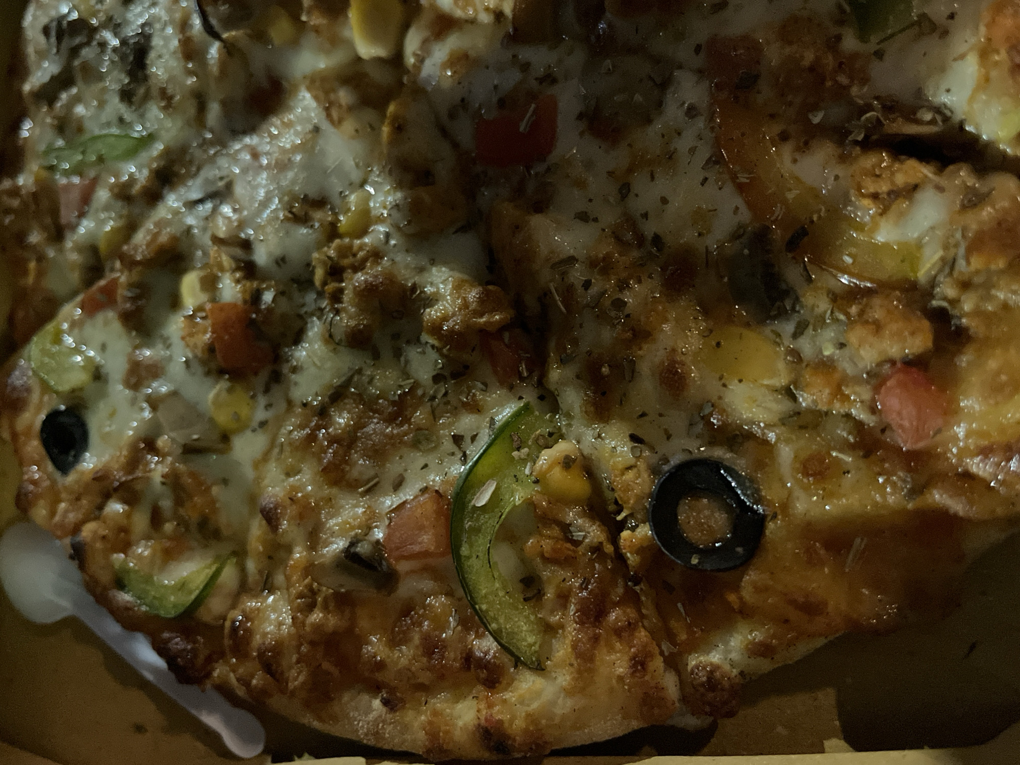 A close-up shot depicts a partially consumed pizza in its cardboard container. Toppings include melted cheese, olives, corn, green bell peppers, tomatoes, and meat. The image was taken indoors in Dhaka, Bangladesh.