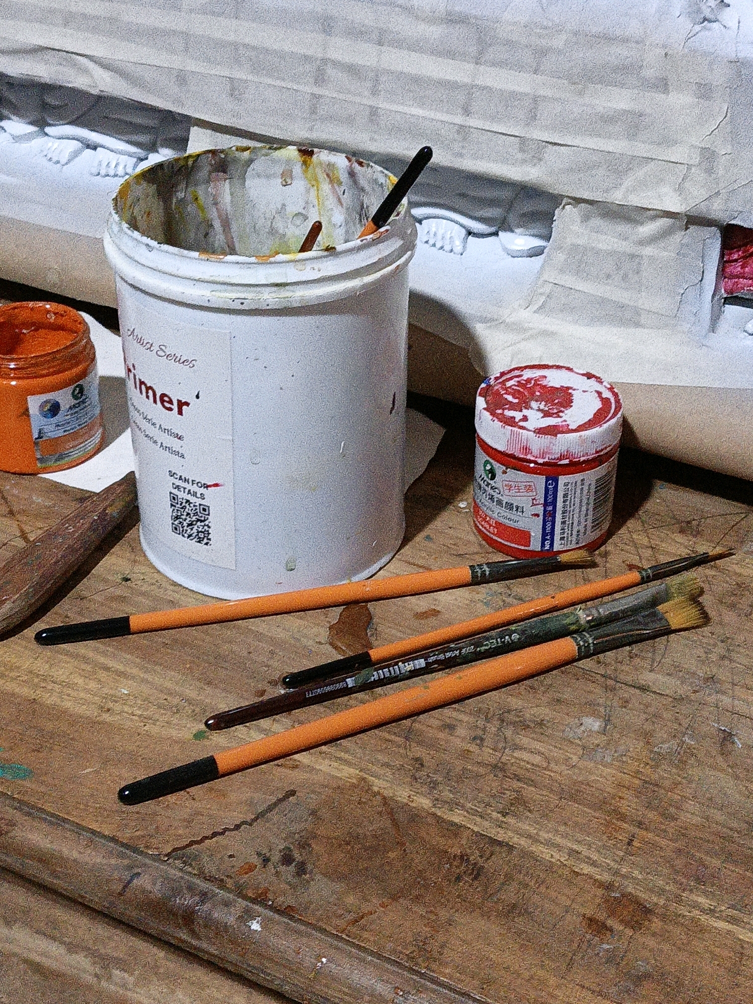 An artist's workstation is depicted in this overhead shot, featuring various art supplies. The scene is set on a worn wooden surface, potentially a table or workbench, stained with paint splatters. In the background, a white object with tape is visible, partially obscuring what appears to be a sculpture or detailed model, possibly depicting human feet.
Dominating the center is a large white bucket labeled "Primer" with "Artist Series" printed above it. The inside of the bucket is stained with dried paint in shades of orange, yellow, and red, with a black-tipped paintbrush resting inside. To the left of the bucket, a small orange jar of paint with a label is present. To the right, a red-lidded jar, also containing red paint, is positioned.
Scattered across the wooden surface are several paintbrushes with orange handles and black tips. Some have visible paint on their bristles. The lighting suggests an indoor environment, possibly a studio. The image does not provide any specific location details for Cikal, Indonesia.