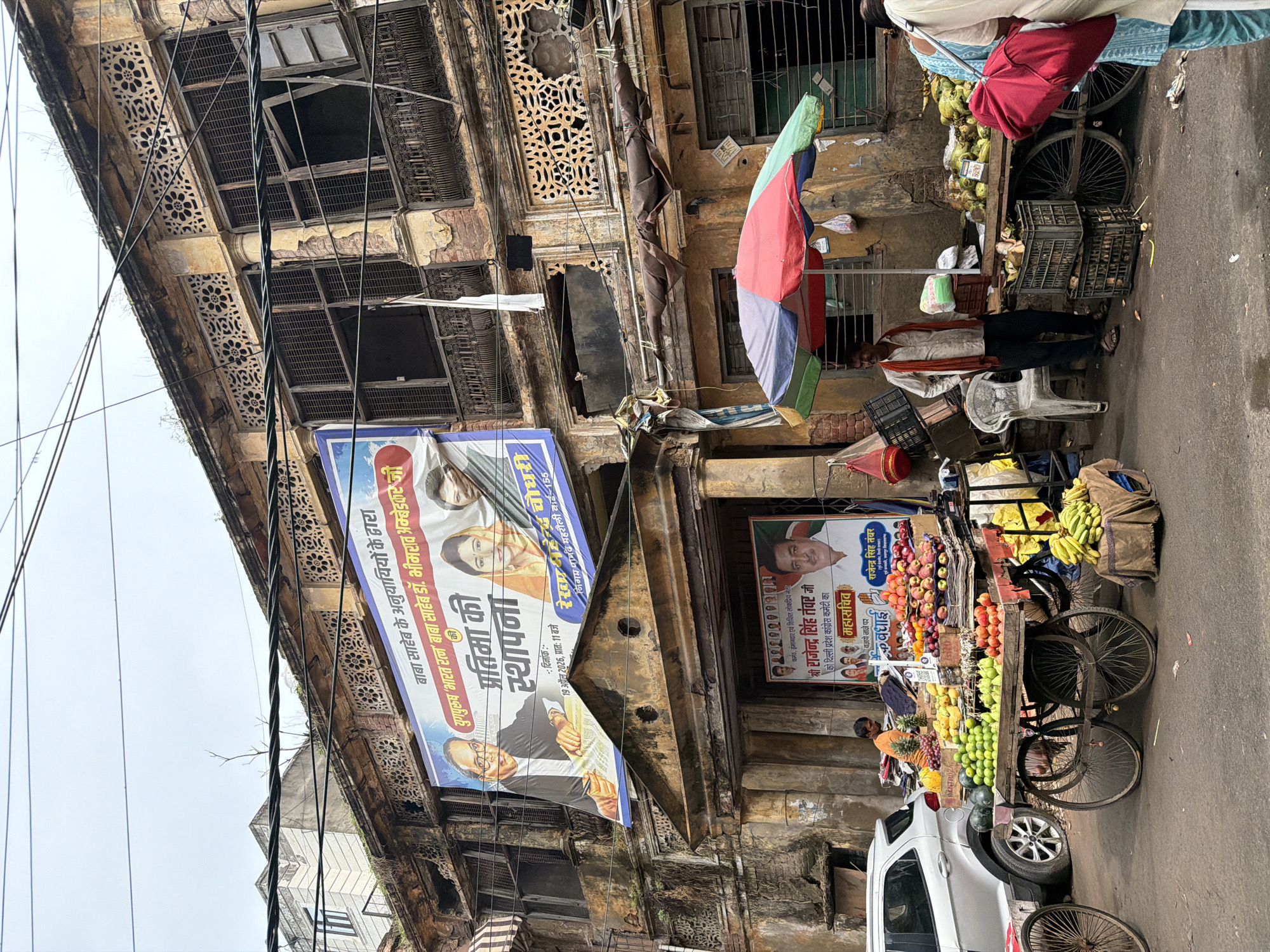 The image depicts a street scene in Delhi, India. In the foreground, a fruit vendor is selling his produce from a cart laden with various fruits, including watermelons, limes, apples, and bananas. Several people are visible around the stall, including the vendor and a customer. Behind the fruit stall, a building with a traditional Indian architectural style stands, featuring ornate balconies and decorative stonework. A large banner is displayed on the building's facade, likely advertising a political event or candidate. The street appears to be a busy marketplace, with other stalls and shops visible in the background. The overall atmosphere is that of a bustling urban environment in Delhi.