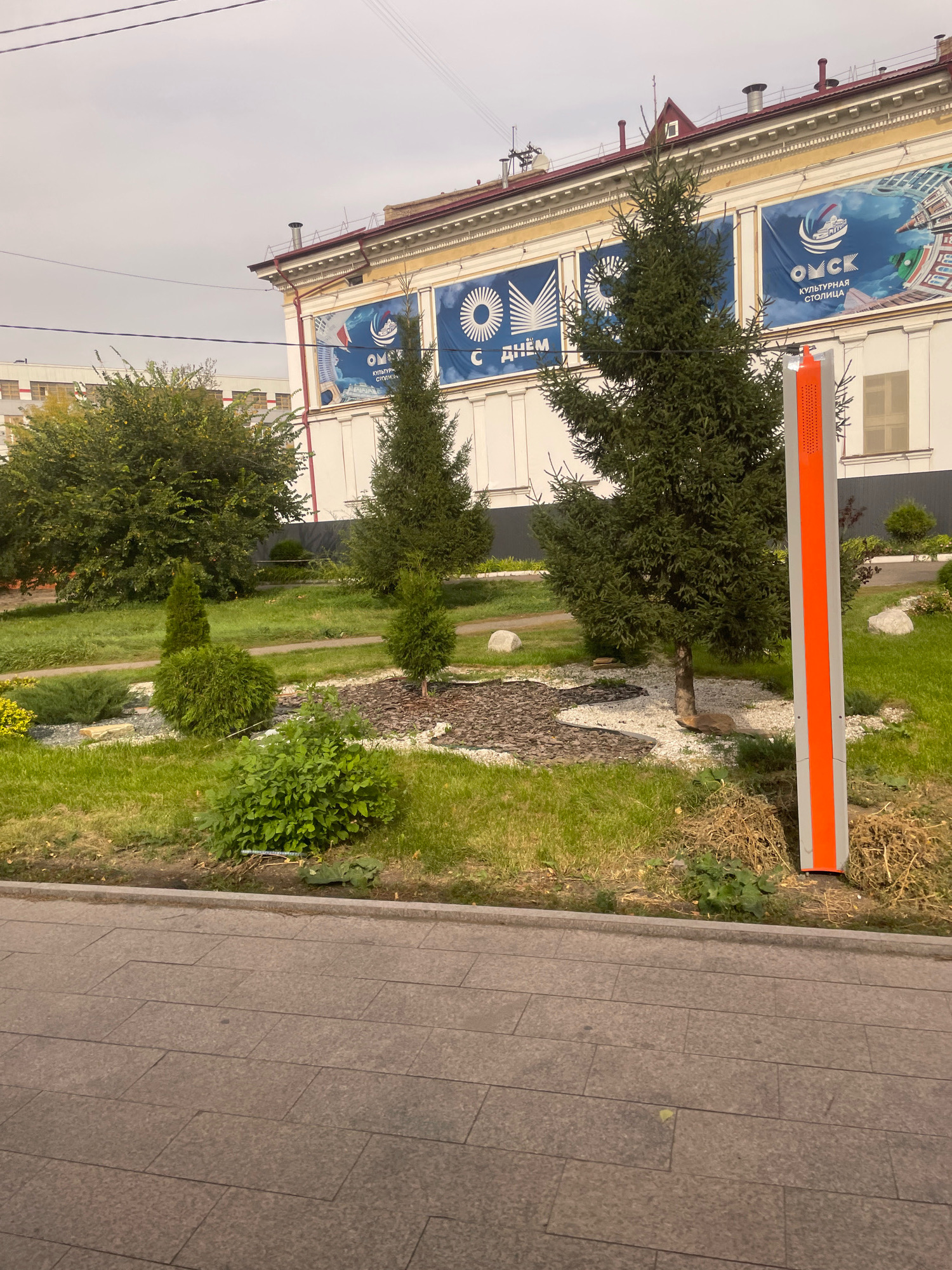 The image shows a street scene in Omsk, Russia. In the foreground, there is a sidewalk made of large, grey stone tiles. Adjacent to the sidewalk is a patch of grass with a decorative garden bed. The garden bed contains a variety of evergreen shrubs and trees, some of which are trimmed into geometric shapes. There is also a tall, orange and grey rectangular sign post on the right side of the image. In the background, a building with a white facade is visible. The building has large banners displayed on its exterior, featuring blue and white graphics and text. The banners appear to be promotional or informational, with one clearly showing the word "ОМСК" (Omsk) and "КУЛЬТУРНАЯ СТОЛИЦА" (Cultural Capital). The sky is overcast.