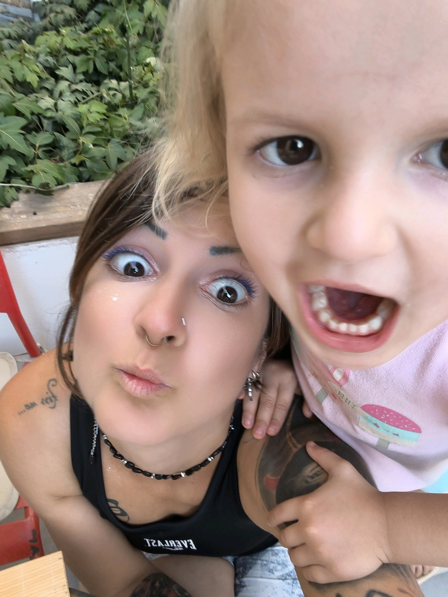 In a close-up, selfie-style photograph taken outdoors, likely in the Lachish Regional Council, Israel, two individuals, a woman and a young child, are prominently featured. The woman, positioned in the foreground to the left, has her eyes wide open and mouth puckered in a playful expression. She is wearing a black tank top with a beaded necklace and has visible tattoos on her arm and shoulder. Her eye makeup includes bright blue mascara. The child, to the right and slightly behind, has their face zoomed in, with their mouth open and teeth visible. They appear to be wearing a pink t-shirt with a graphic print. The background consists of lush green foliage and some wooden elements, suggesting an outdoor setting. The lighting is bright, indicating daylight.
