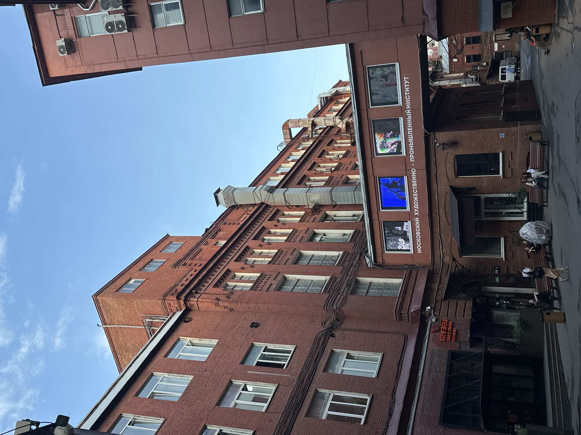 The image shows a courtyard surrounded by red brick buildings in Moscow, Russia. A pedestrian bridge with large digital screens displaying colorful images spans across the courtyard, connecting two of the buildings. The text on the bridge reads "МОСКОВСКИЙ ХУДОЖЕСТВЕННО-ПРОМЫШЛЕННЫЙ ИНСТИТУТ" (Moscow Art-Industrial Institute). On the ground floor of one building, there is a sign that says "МУЗЕЙ" (Museum). Several people are seated on benches in the courtyard, and one person is walking. The sky is blue with some clouds. Air conditioning units are visible on the exterior walls of some buildings, and various pipes and architectural elements are present. The overall scene depicts a vibrant urban setting, possibly a university campus or a cultural complex, with a mix of architectural styles and contemporary elements.