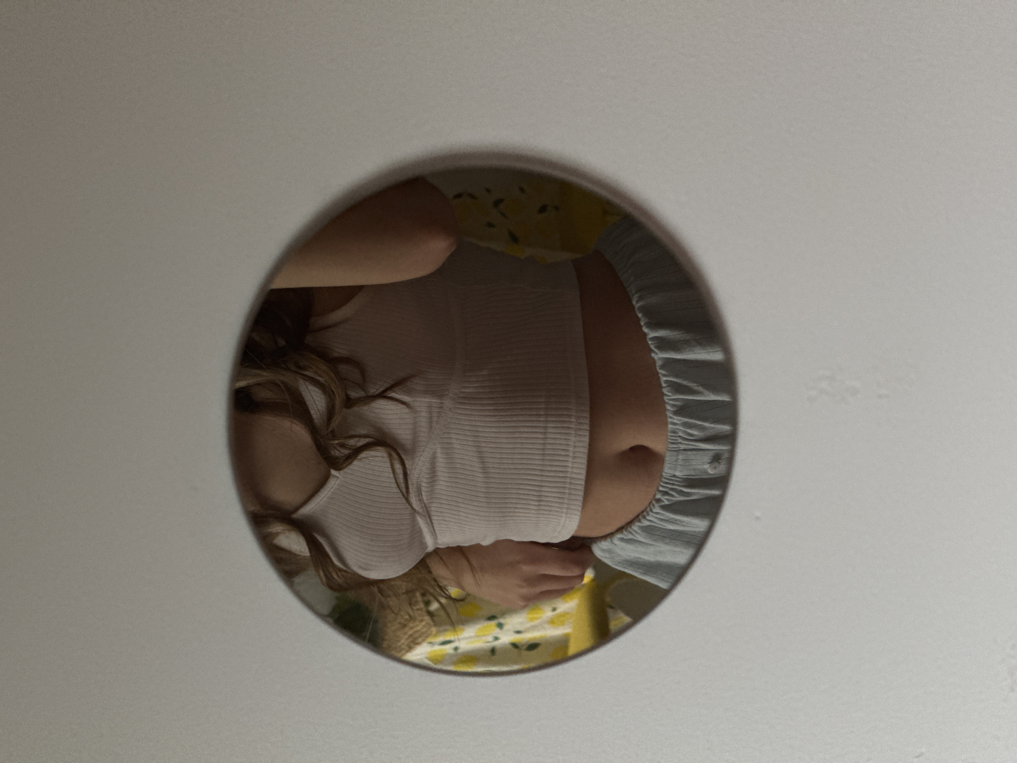 A round mirror hangs on a white wall, reflecting a person in a white tank top and light blue pajama bottoms. The person's face is partially visible with blonde hair. They are in a room with a yellow and white patterned bedspread.