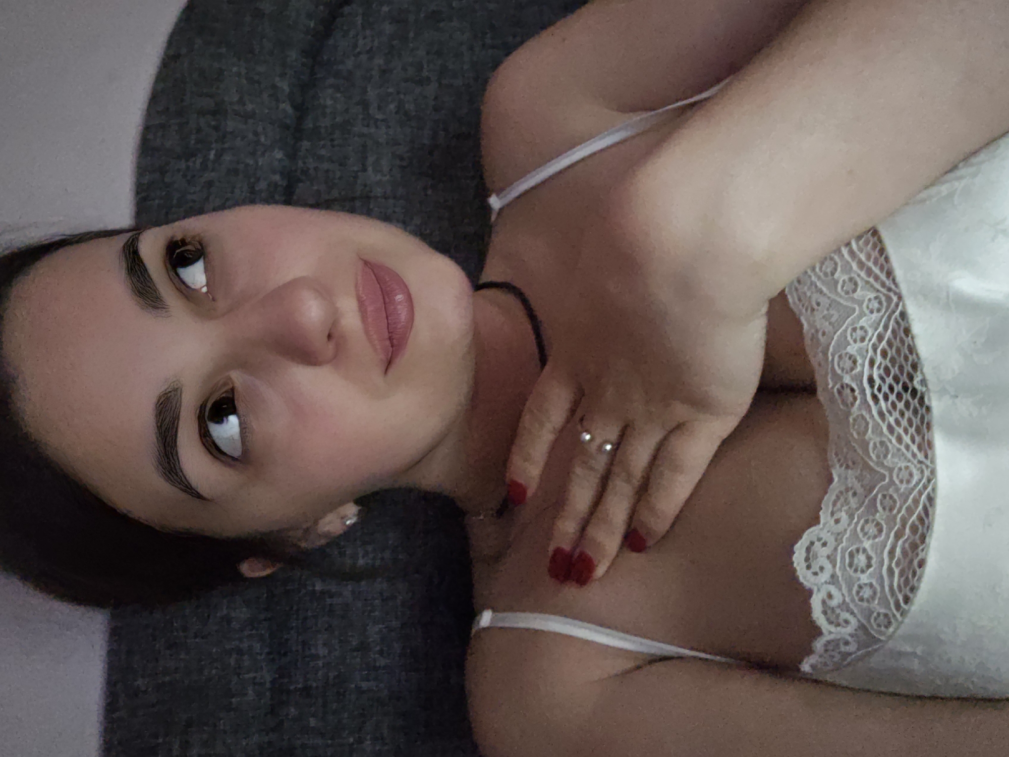 A young woman is lying on a gray sofa in Petropavlivska Borshchahivka, Ukraine. She is wearing a white lace-trimmed camisole. Her right hand is resting on her chest, with red-painted nails and a silver ring on her index finger. Her left hand is not visible. The woman has dark hair pulled back, dark eyebrows, and is looking to her right with a thoughtful expression. The lighting suggests it is nighttime or a dimly lit indoor space.