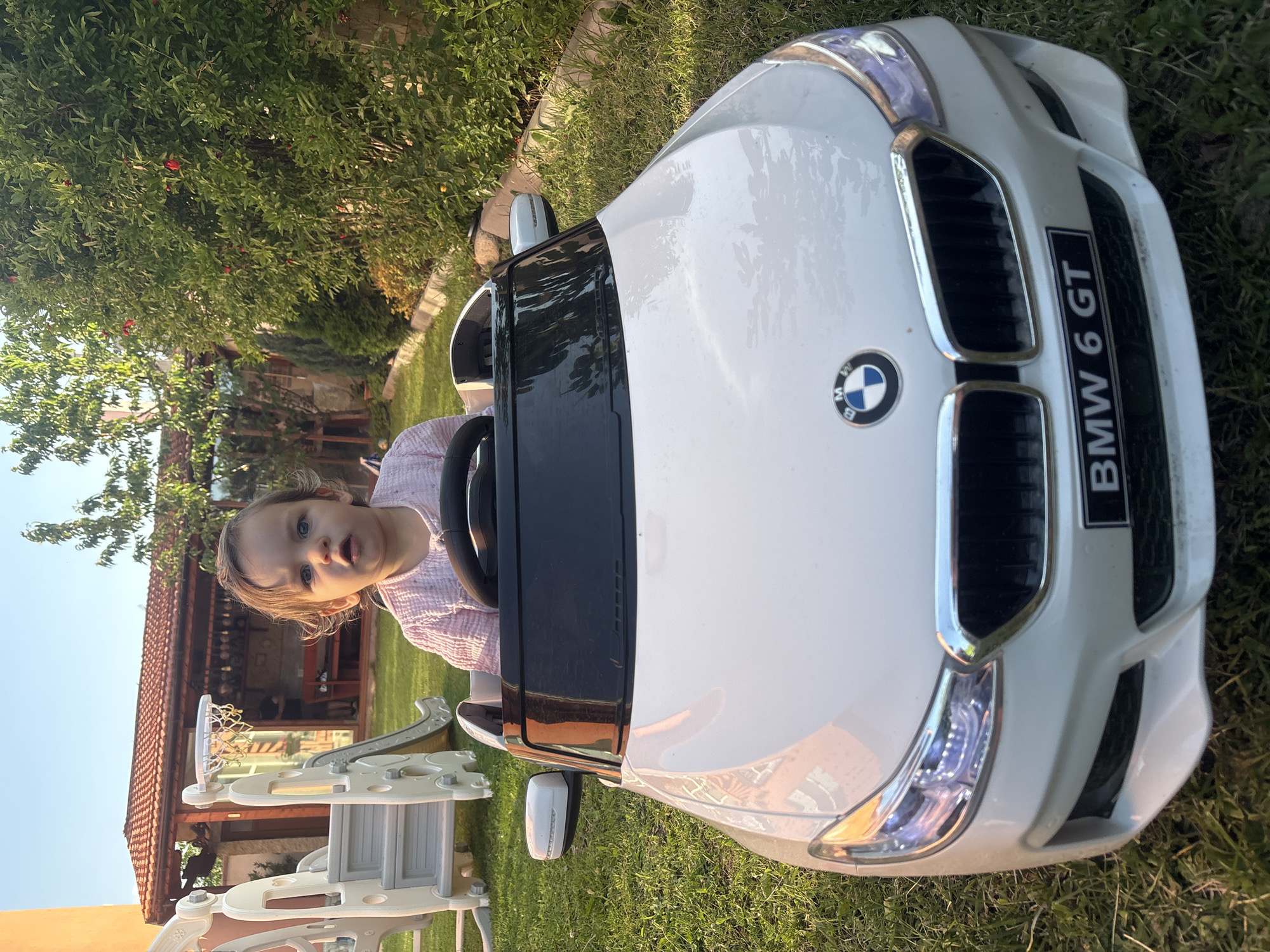 In Strelcha, Bulgaria, a young child, possibly a toddler, sits in the driver's seat of a white toy BMW 6 GT car on a green grassy lawn. The child wears a pink, textured garment and has a curious expression. The car is positioned in a backyard, with a multi-level children's slide and a small basketball hoop visible in the background, to the left. Beyond the play equipment, a covered outdoor seating area with a tiled roof is present. Lush green trees and bushes, some with small red fruits, flank the scene, suggesting a garden setting. The lighting indicates it is late afternoon or early evening. The overall impression is of a playful outdoor environment.