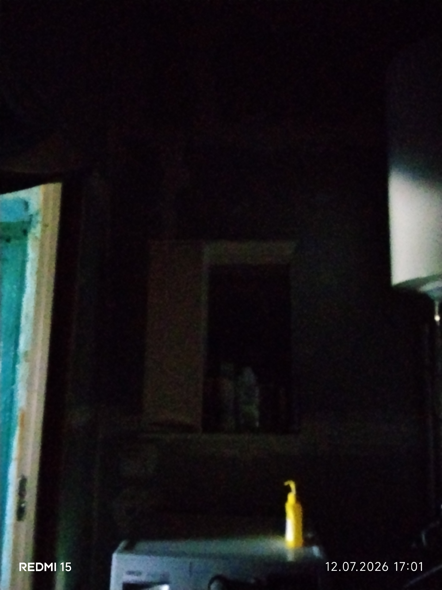 The image shows a dimly lit interior scene, possibly a room. On the left, a portion of a doorway or frame with teal and white colors is visible. In the center, there is a small, dark opening, possibly a window or alcove, within a dark wall. Below this opening, on a dark surface, sits a bright yellow pump bottle. In the lower right corner of the image, a timestamp reads "12.07.2026 17:01". In the lower left corner, text reads "REDMI 15". The overall impression is one of low light conditions. The location of Айет, Kazakhstan is not discernible from the image itself.
