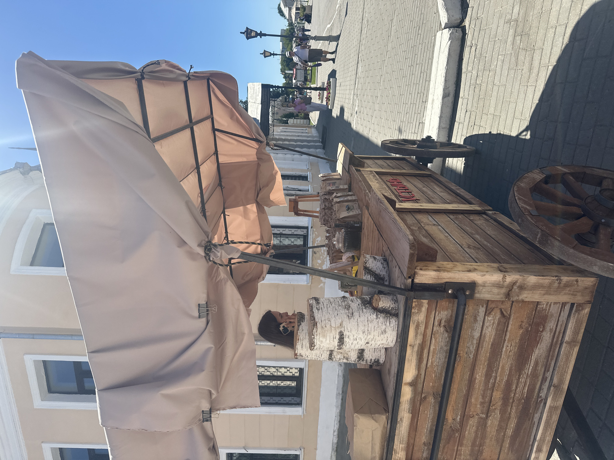A woman is standing behind a wooden cart on wheels, holding a phone to her ear. The cart is covered by a light-colored canvas awning and displays several bags of goods and decorative birch logs. The scene appears to be an outdoor market or street vendor setup on a paved sidewalk. In the background, a building with light-colored walls and windows is visible, along with other people walking and street lamps. The overall setting suggests a sunny day in a historic urban environment, potentially the city of Kolomna, Russia, given the architectural style and the presence of a street vendor.