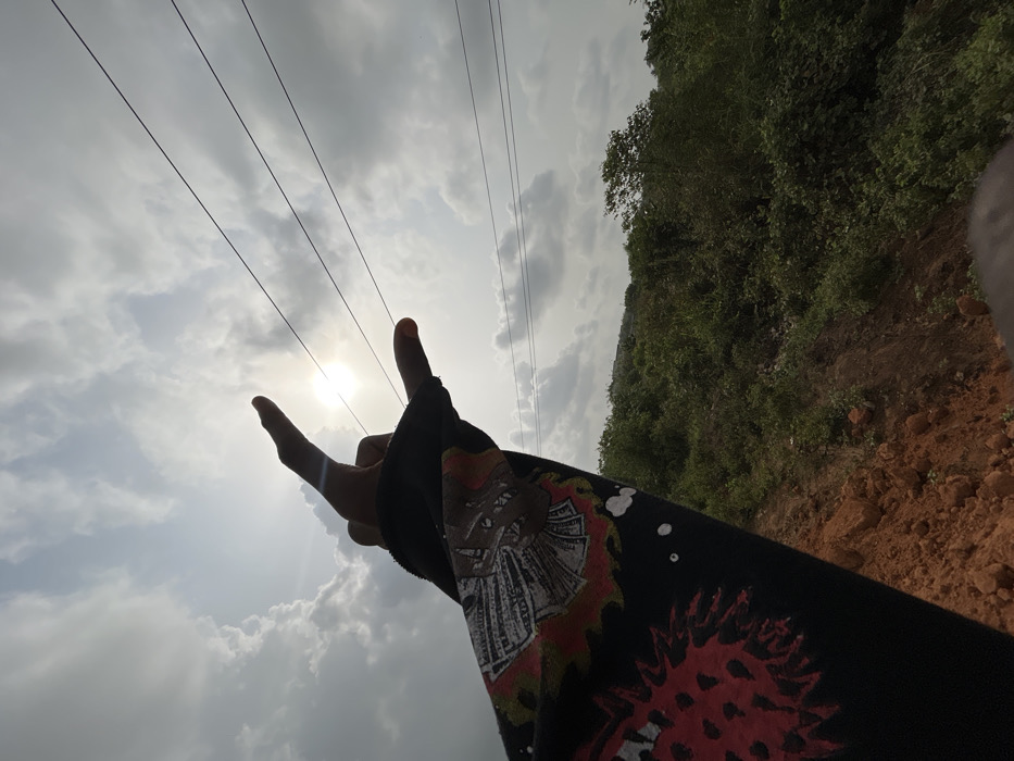 The image depicts a close-up, low-angle perspective of a human left arm and hand, silhouetted against a cloudy sky with the sun visible. The hand, positioned in the upper left quadrant, forms a gesture with the thumb, index, and pinky fingers extended upwards, while the middle and ring fingers are curled into the palm. The wrist and forearm are covered by a dark, long-sleeved garment featuring multiple printed graphics; one visible graphic includes a stylized head with what appears to be wings or currency notes, and further down the sleeve, a prominent red and orange spiky design resembling a virus or monster.
In the midground, extending diagonally across the upper right portion of the frame, are three dark, parallel power lines. The background consists of an outdoor landscape under an overcast sky with a bright, partially obscured sun. Below the horizon line, the terrain includes a sloping bank of reddish-brown exposed soil, transitioning into dense green vegetation and bushes that cover hillsides. A faint outline of what could be a distant structure or ridge is visible on the far left horizon. The scene is located in the city of Ilishan, Nigeria.