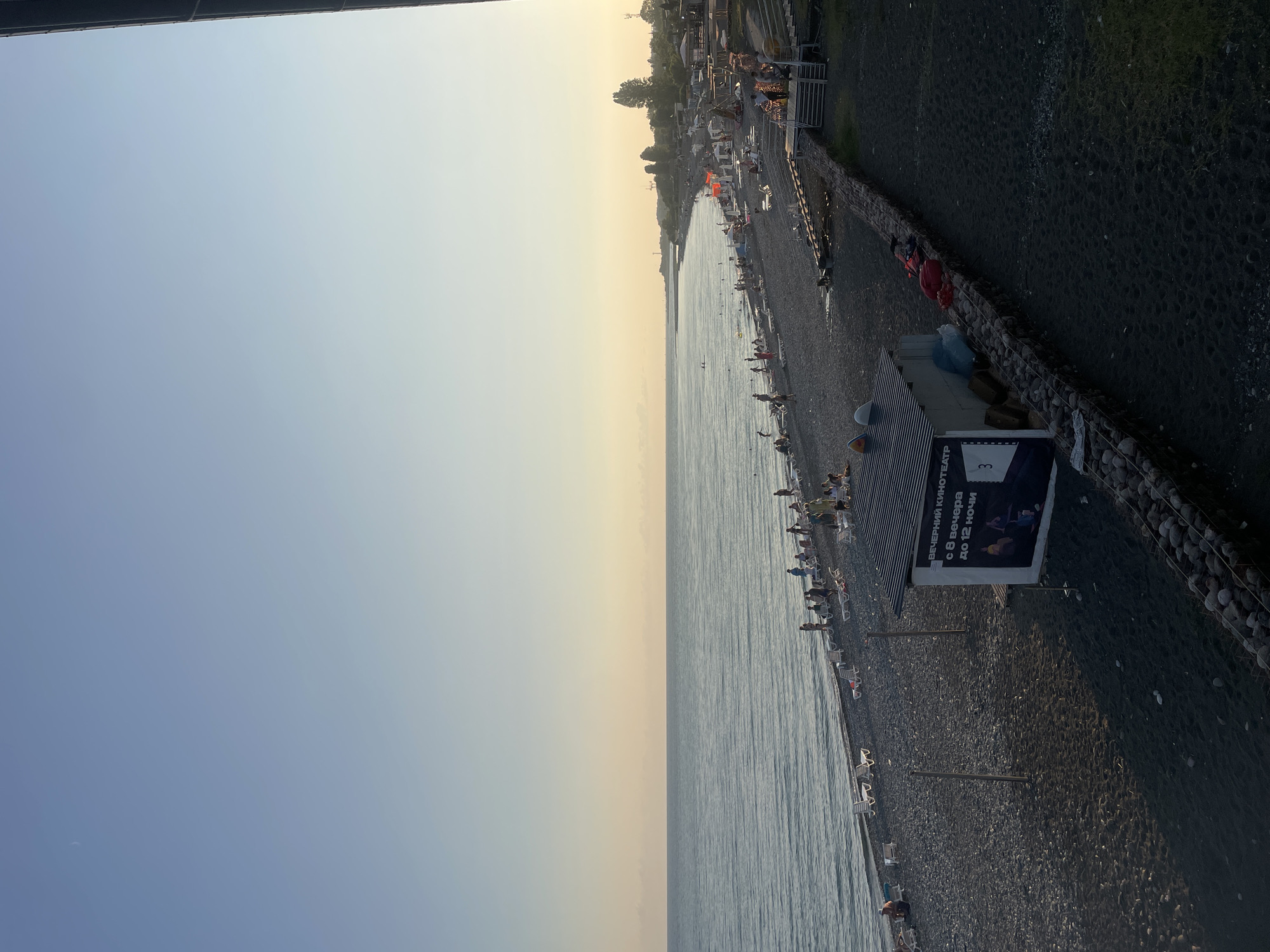 The image depicts a wide view of a crowded pebble beach and the adjacent sea in Giachrypsh, Abkhazia. The sky is a soft gradient of pale blue to light yellow, indicating either early morning or late afternoon. A faint crescent moon is visible in the upper left portion of the sky. The sea is calm with gentle waves lapping at the shore.
The beach is covered in dark grey pebbles and is dotted with numerous sunbathers, many of whom are seated on lounge chairs arranged in rows. People are seen walking along the shore, some are wading in the water, and others are gathered in small groups. The beach is lined with umbrellas and small structures.
In the foreground, to the right of the image, there is a dark, sandy area. A low wall constructed of large stones separates this area from the pebble beach. On the pebble beach, near the center of the image, is a small, shed-like structure with a striped roof and a sign that reads "ВЕЧЕРНИЙ КИНОТЕАТР" which translates to "Evening Cinema." The sign also indicates that the cinema operates from 8 PM to 12 AM. Several objects, including red and blue bags, are placed near this structure.
Further down the beach, there are more structures, some appearing to be covered seating areas or cafes, and a set of stairs leading up to a higher level. Trees are visible in the distance along the coastline, and the overall atmosphere suggests a popular tourist destination during the day.
