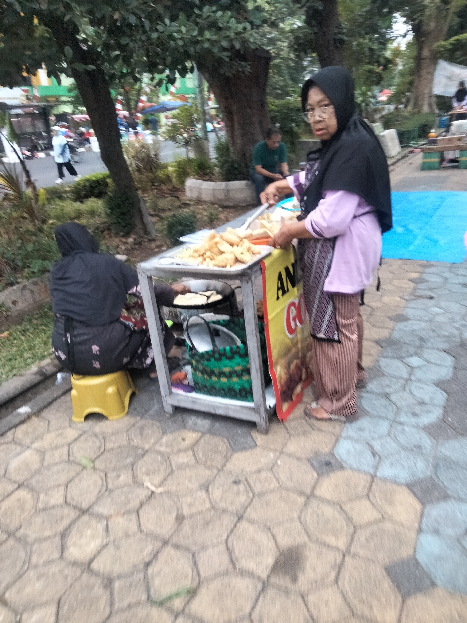 In Surakarta, Indonesia, two women are seen operating a food stall in an outdoor urban setting. A woman in the foreground, wearing a black hijab and a light purple top with striped brown pants, stands behind a mobile food cart. She is holding a pair of tongs and appears to be serving food from a tray of fried items. The cart has a yellow and red sign with the text "AN" and "GO" visible. To the left of the cart, another woman, dressed in a black hijab and a dark floral patterned dress, sits on a yellow stool. She is facing away from the camera, towards the cart. The food cart contains a pan with oil, a basket, and other items. The ground is paved with hexagonal tiles in varying shades of beige and gray. In the background, trees, other stalls, and people can be observed, suggesting a public area or market. A bright blue tarp is visible on the right side of the frame.