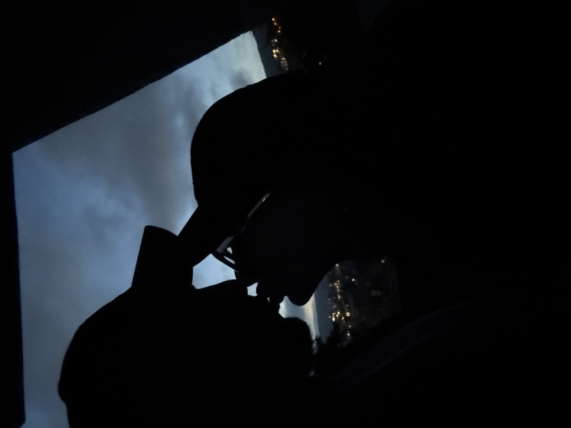 A silhouette of a person wearing glasses and a hat is seen against a window. The view through the window suggests a night scene with scattered lights, possibly indicating a cityscape. Based on the prompt, this is purportedly located in Varna, Bulgaria. The person is looking down, seemingly engaged in some activity obscured by the darkness.