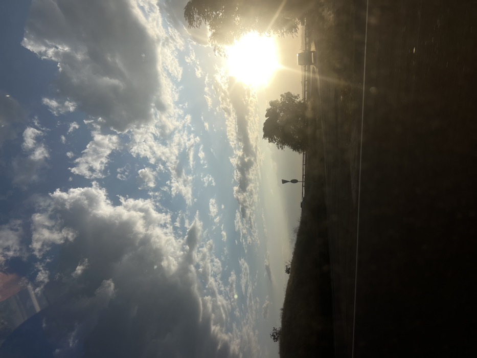 A bright sun shines through a cloudy sky over a roadside scene. To the right, a paved road stretches out beside a grassy verge with trees and a reflective sign. The sun's glare overwhelms part of the view, highlighting trees and the edge of the road. The focus is on the sky filled with scattered clouds, creating a bright, airy atmosphere.