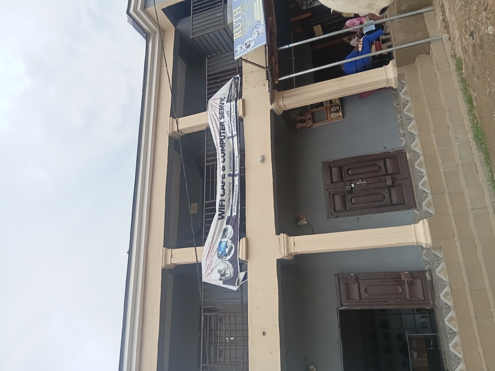 The image shows the exterior of a multi-story building. The building is painted a light beige color and has several windows and doorways. A banner hangs from the building, advertising "WIFI CAFE & COMPUTER SERVICE". The banner lists services such as "Internet Cafe", "Computer Training", "Lamination", "Photocopying", and "Printing". A phone number is also visible on the banner. A smaller sign above the banner reads "RUTH".
On the upper floor of the building, people are visible through an open area. One person is wearing a blue top and appears to be seated. The sky is overcast, suggesting it might be a cloudy day. The lower part of the image shows a staircase leading up to the building. The scene appears to be an urban or semi-urban commercial area in Jalingo, Nigeria, given the context. The overall impression is of a business establishment providing various services to the public.
