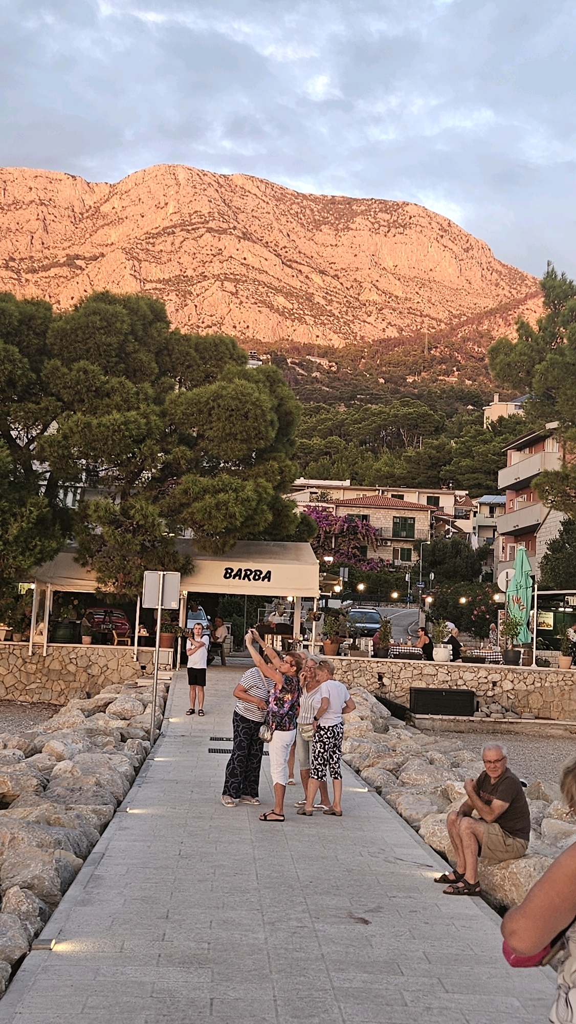 In Općina Tučepi, Croatia, a group of four women are taking a selfie on a paved walkway. The women are positioned centrally in the frame, with one woman holding up her phone to capture the photo. They are casually dressed, with varying patterns and colors on their clothing. Behind them, a restaurant or cafe with the word "BARBA" visible on its awning is situated. The walkway is lined with large rocks, and beyond that, there is a beach area. In the background, lush green trees and buildings are visible, leading up to a large, rocky mountain illuminated by warm, sunset-like lighting. To the right of the walkway, a man is seated on a rock, observing the scene. The sky above is partly cloudy, with a gradient of light blue and gray.