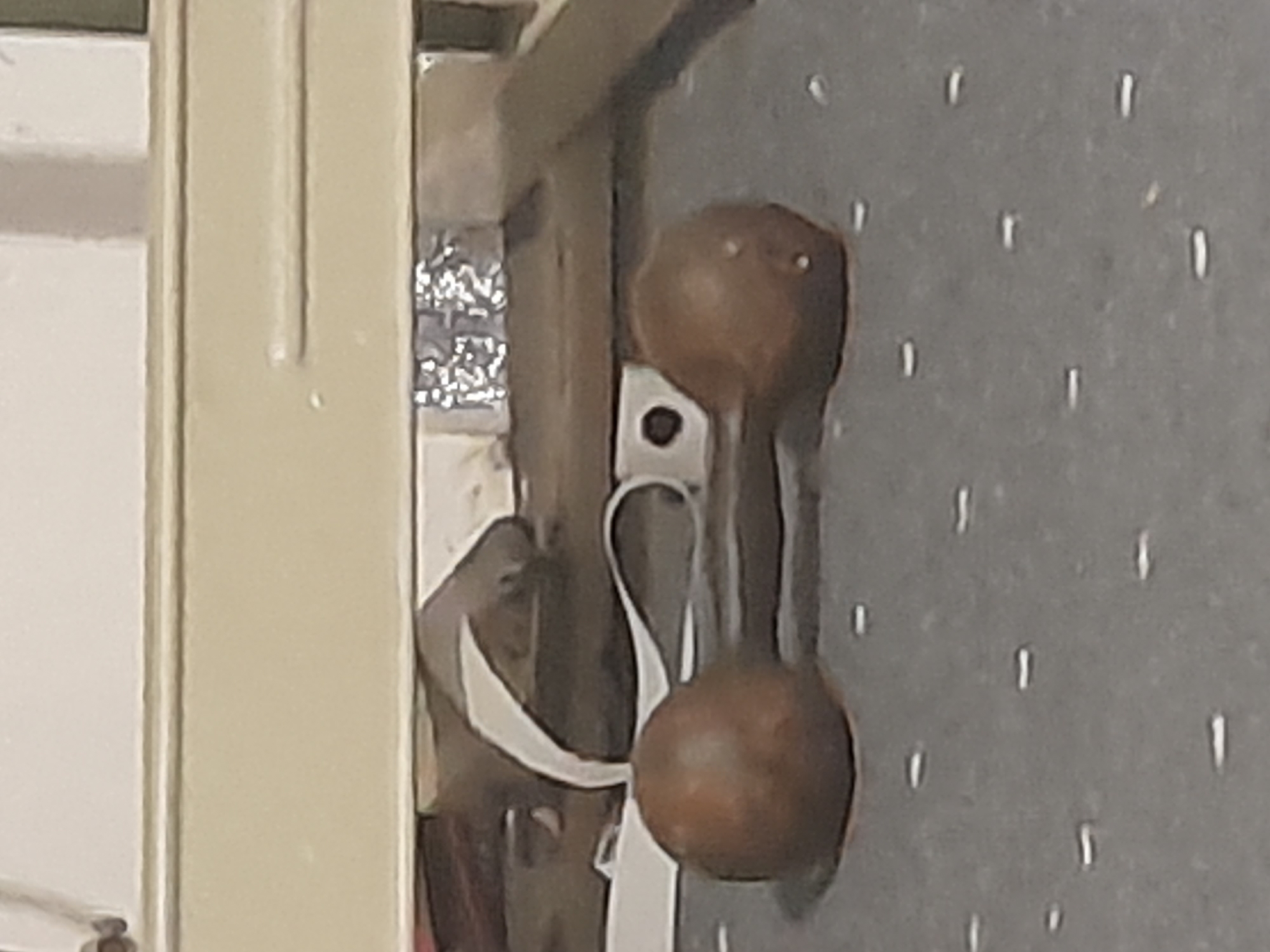 The image shows a close-up view of a pair of small, brown dumbbells lying on a gray, speckled surface. A white strap or ribbon is visible behind the dumbbells, partially obscuring a wooden surface. The background is out of focus but appears to be a room with a light-colored wall and a ledge or shelf. There are no people or animals visible in the image. The setting is a room in Кароол, Kyrgyzstan.