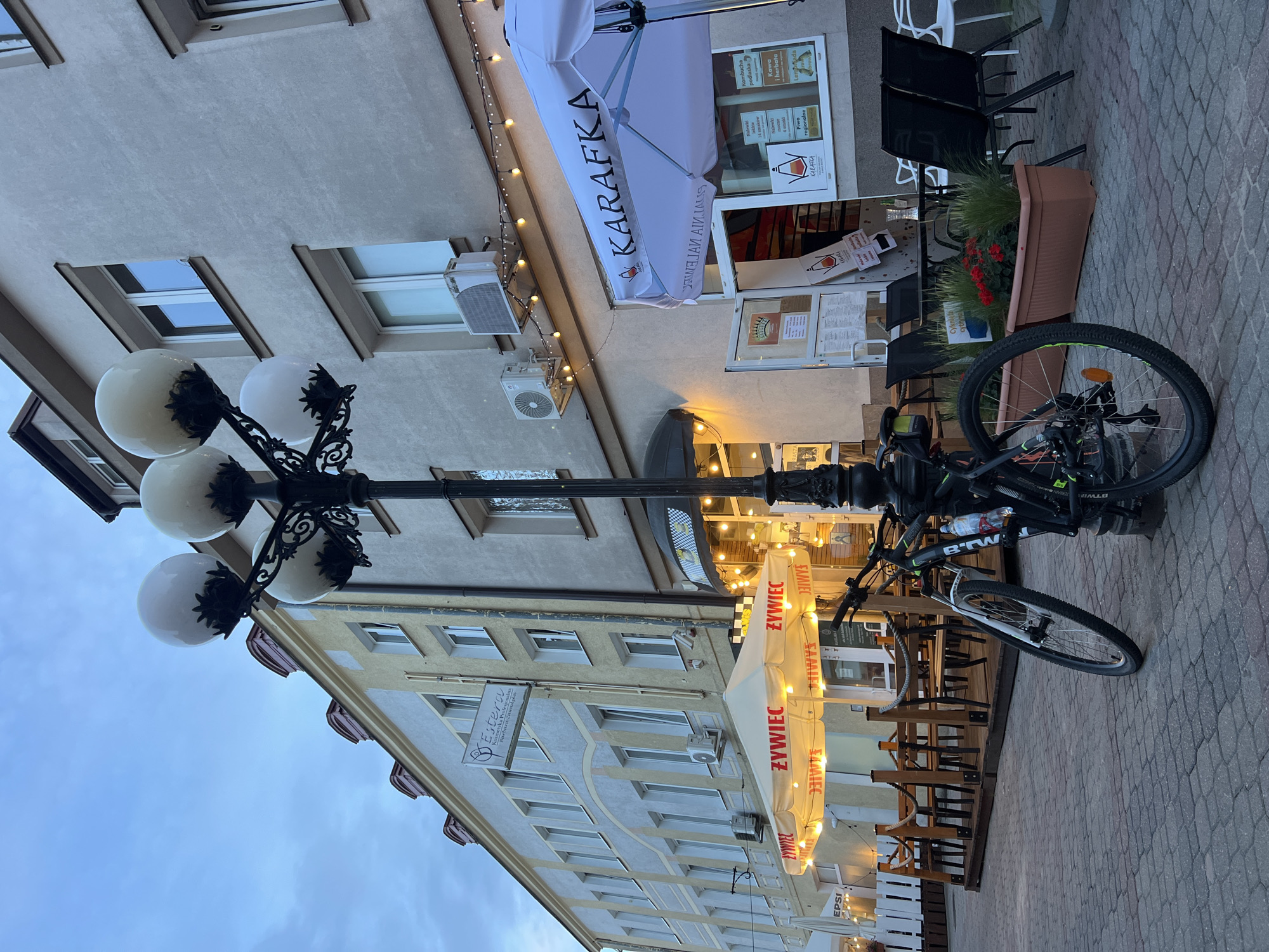 The image depicts an outdoor seating area of a cafe or restaurant on a cobblestone street in Białystok, Poland. The time of day appears to be dusk, with the sky a muted blue and artificial lights beginning to illuminate the scene. In the foreground, a bicycle is parked next to a decorative lamppost with multiple globes. The lamppost is ornate and made of black metal.
The cafe's facade is visible, featuring a white umbrella with the logo "KARAFKA" in large black letters. String lights are draped across the building above the entrance. The cafe has an outdoor seating area with wooden tables and chairs, some of which are occupied by patrons. A portion of the building on the left has a sign that reads "Estera Kosmetyka Profesjonalna" indicating a beauty salon.
The overall atmosphere is calm and inviting, suggesting a pleasant evening in a European city. The architecture is typical of urban European settings, with multi-story buildings lining the street. The cobblestone pavement and the outdoor seating arrangement are characteristic of many European towns and cities.