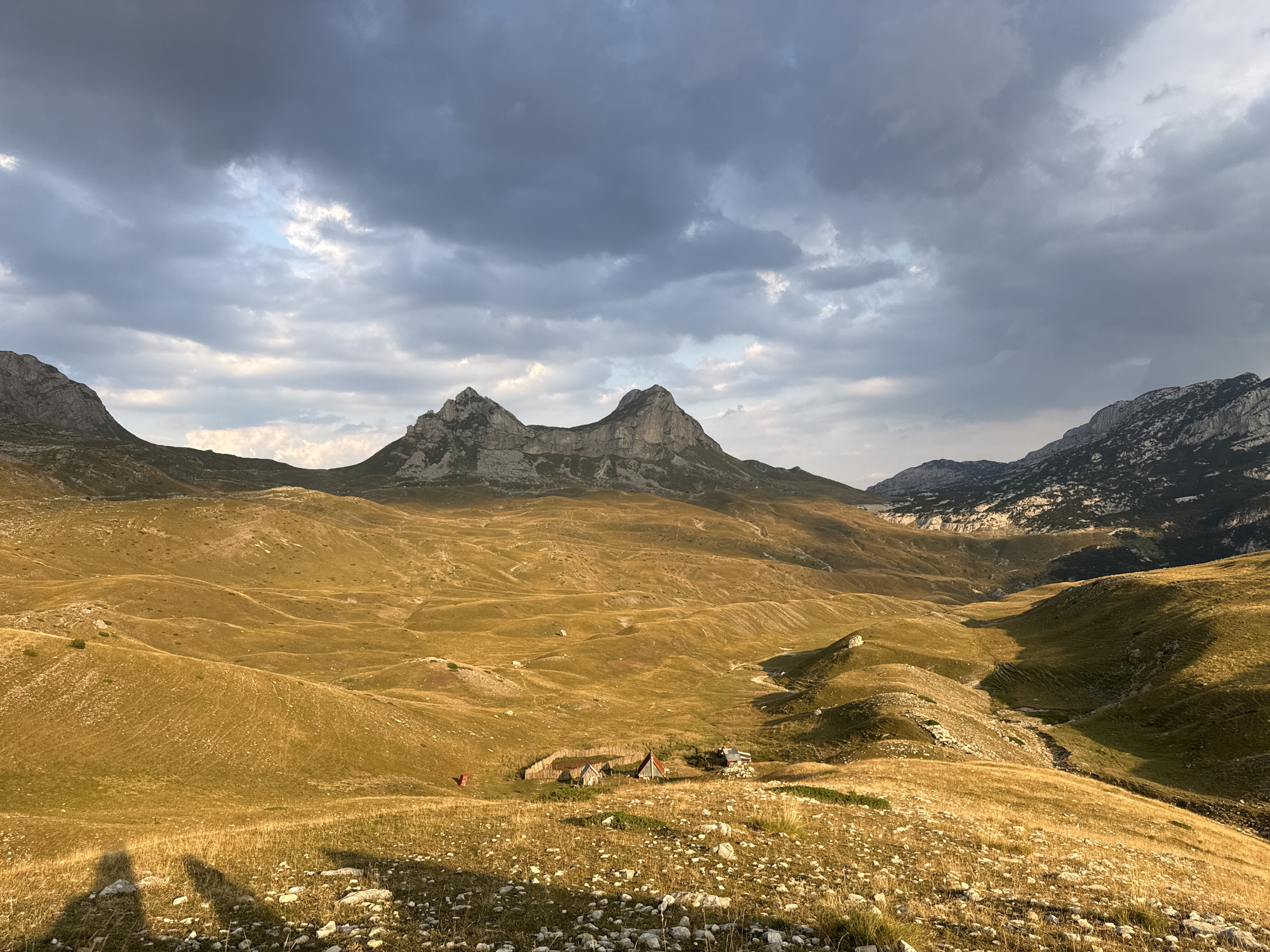 The image depicts a mountainous landscape in Dubrovsko, Montenegro. The foreground is dominated by rolling hills covered in dry, golden-brown grass and scattered white rocks. Several small, rustic structures, resembling cabins or shelters, are clustered in a valley in the middle ground. A red object, possibly a tent or a piece of equipment, is visible to the left of these structures. Shadows of two figures are cast on the ground in the lower left corner of the image. In the background, majestic, rugged mountains rise against a dramatic sky filled with dark, brooding clouds interspersed with patches of lighter sky. The overall impression is of a remote, wild, and expansive natural environment.