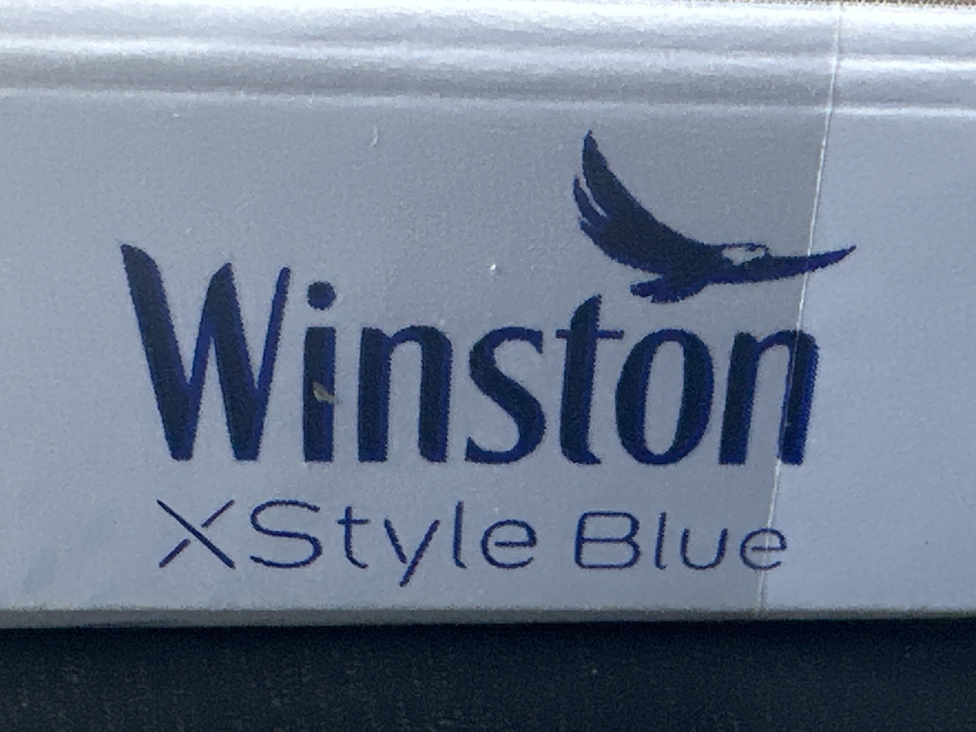 The image shows a close-up of a blue and white cigarette pack. The brand name "Winston" is printed in large, dark blue letters. Below that, in smaller, dark blue letters, is "XStyle Blue". A silhouette of a bird in flight is positioned to the right of "Winston". The background appears to be a textured surface, possibly a table or a shelf. The image quality is somewhat grainy, with some minor imperfections on the surface of the pack. The location of the image in Putilkovo, Russia, is not discernible from the visual information provided.