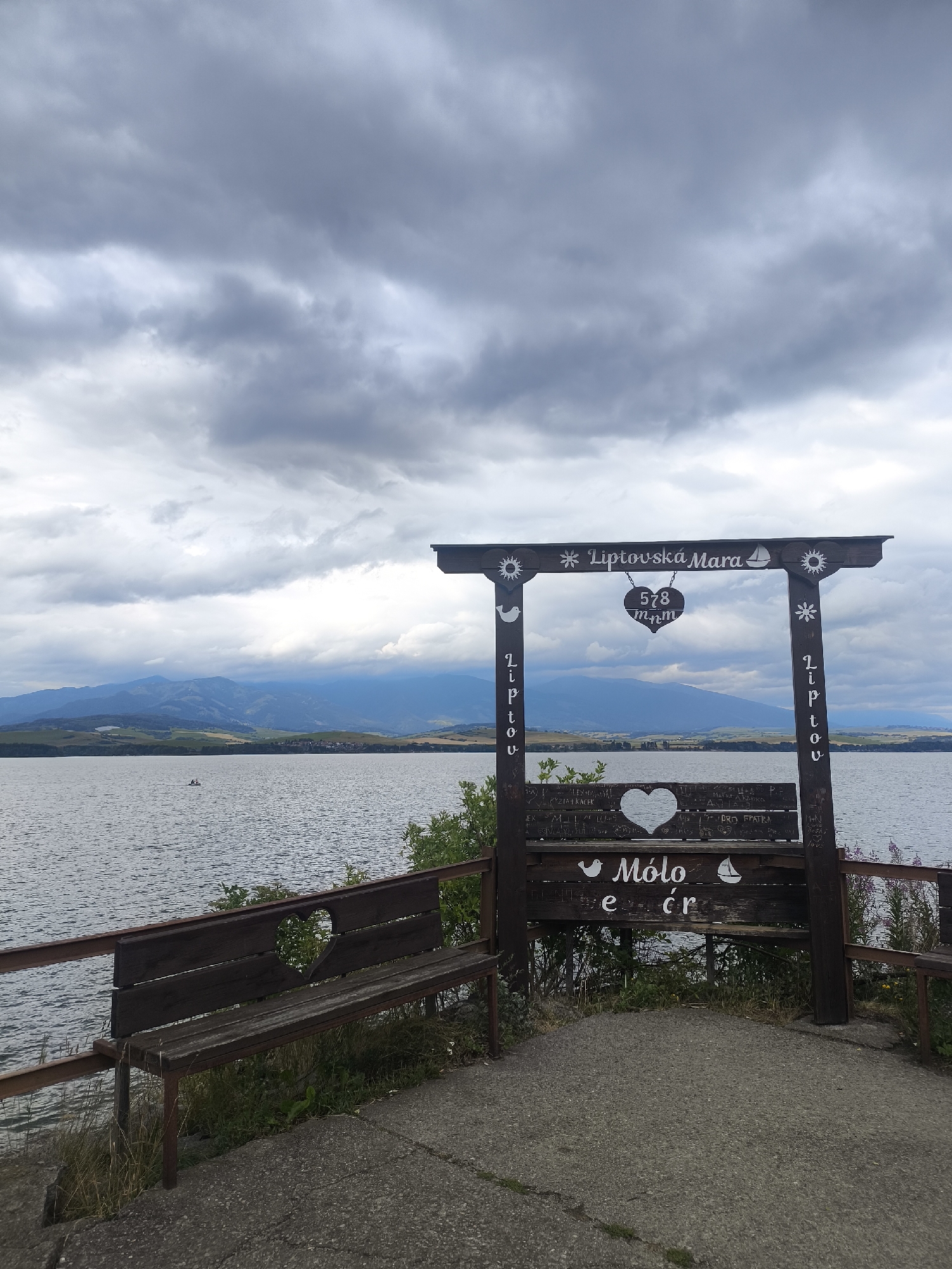 This is a medium-shot, eye-level, outdoor photograph taken on a cloudy day at the Molo pier in Galovany, Slovakia, a location on the Liptovská Mara reservoir. In the foreground, a rustic wooden bench with a heart-shaped cutout is positioned on a paved area. The bench is part of a larger wooden archway structure with decorative carvings of a sun, a sailboat, and a bird. Inscriptions are visible on the archway and the bench, including "Liptovská Mara," "578 m n.m." (meters above sea level), and "Molo." The reservoir stretches across the midground, with a distant mountain range visible on the horizon. The water is calm, with a small boat containing two people visible in the distance. The sky is heavily overcast with dramatic clouds.