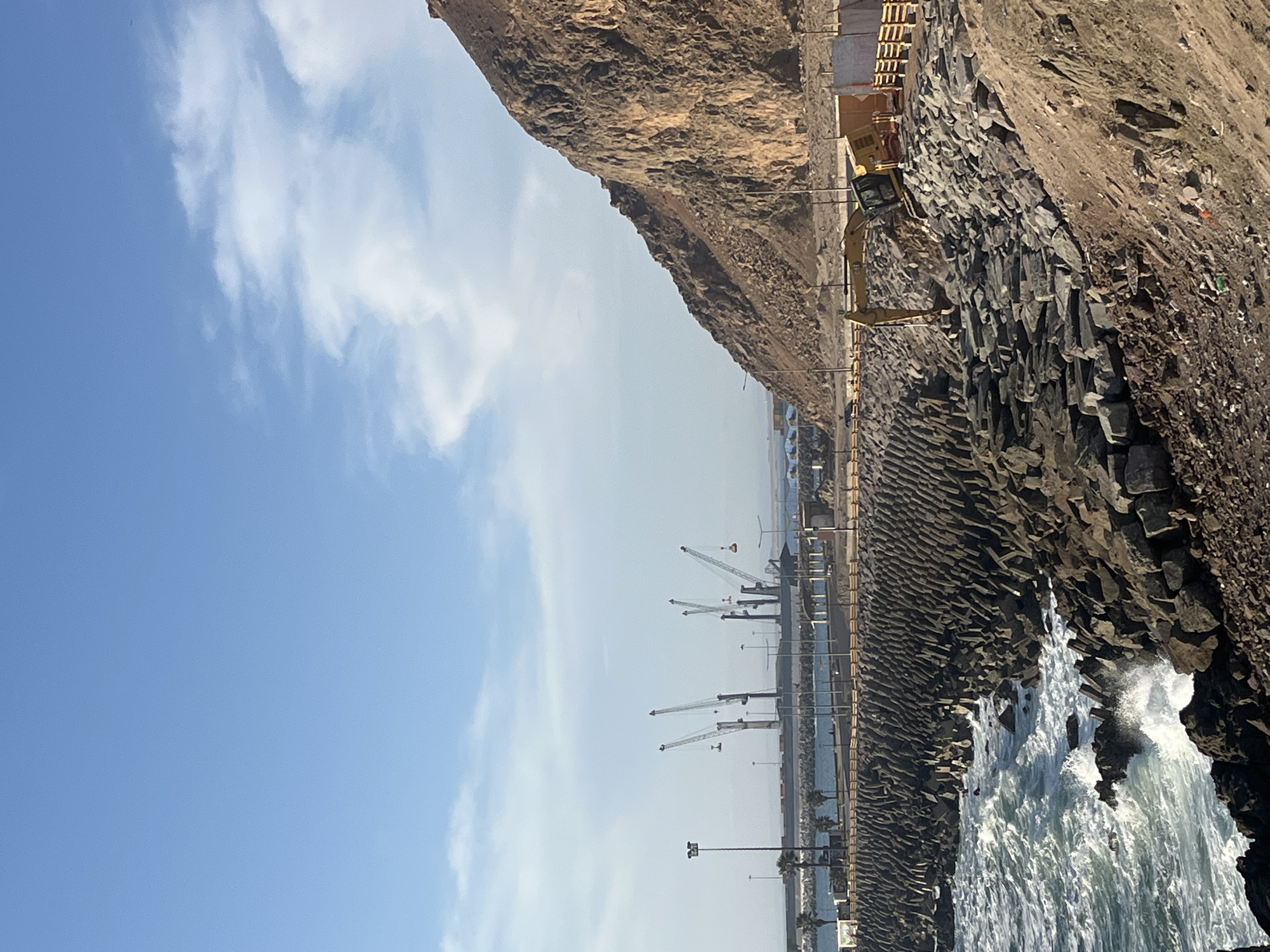 The image shows a coastal scene in Arica, Chile. On the right, there is a rocky cliff face, with waves crashing against the base of the rocks. A line of large, grey, blocky rocks lines the water's edge, likely forming a breakwater or seawall. An excavator sits atop the rocks near some wooden building materials. A harbor with several cranes is visible in the distance. The sky is mostly clear with some scattered clouds.