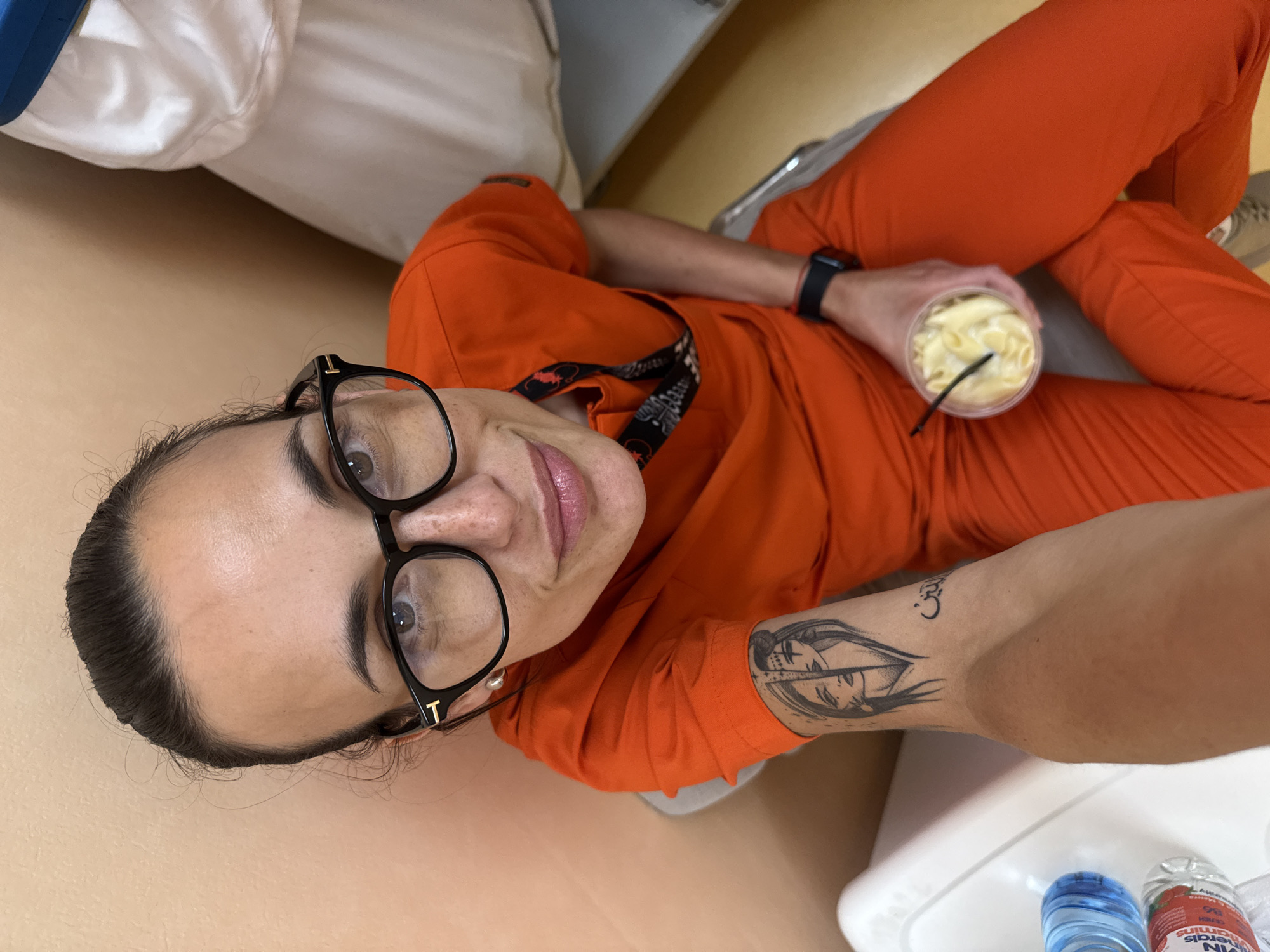 The image captures an adult woman in an indoor setting, possibly a room, from a high-angle perspective, likely a selfie. She is lying or reclining, looking directly at the camera with a slight smile and wide-eyed expression. Her dark hair is pulled back, and she wears black-framed glasses with a small gold 'T' detail on the side.
She is dressed in an orange long-sleeved top and matching orange pants, resembling a uniform or scrubs. A patterned lanyard is visible around her neck, and a black smartwatch is on her left wrist. On her right forearm, she has a tattoo depicting a stylized face or two faces within an eye-like shape, with a small symbol resembling Om/Aum or a stylized '3' above it.
In her right hand, she holds a clear plastic cup containing a light yellow, textured substance, possibly mac and cheese or a similar creamy dish, with a black straw. To her right, on a white surface, are two small plastic bottles; one has a blue cap, and the other, with a red/pink label, shows partially visible text including "VITAMIN WATERS," "MINERALS," "strawberry," and "50 CALORIES."
The background includes a light brown wall or surface and a section of white bedding or a pillow in the upper left. The overall impression is of a casual moment, possibly a break or relaxation. The scene is set indoors; the provided geographical context is Pleven, Bulgaria.