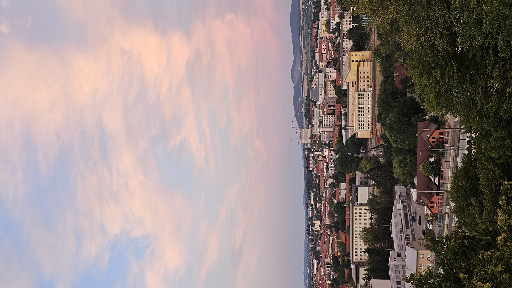 The image captures an aerial view of the city of Prešov, Slovakia, at dusk. The sky is filled with soft, diffused light, painted with streaks of pink and pale blue from the setting sun. Dominating the midground is a sprawling urban landscape characterized by a mix of architectural styles. Red-tiled roofs are prevalent on many buildings, interspersed with modern, multi-story structures, some of which appear to be educational institutions or office buildings. Construction cranes are visible in the distance, suggesting ongoing development within the city. Lush green trees form a dense canopy in the foreground and throughout the cityscape, adding a natural element to the urban environment. The overall impression is one of a serene evening settling over a populated area, with the vibrant colors of the sky setting a peaceful, atmospheric tone.