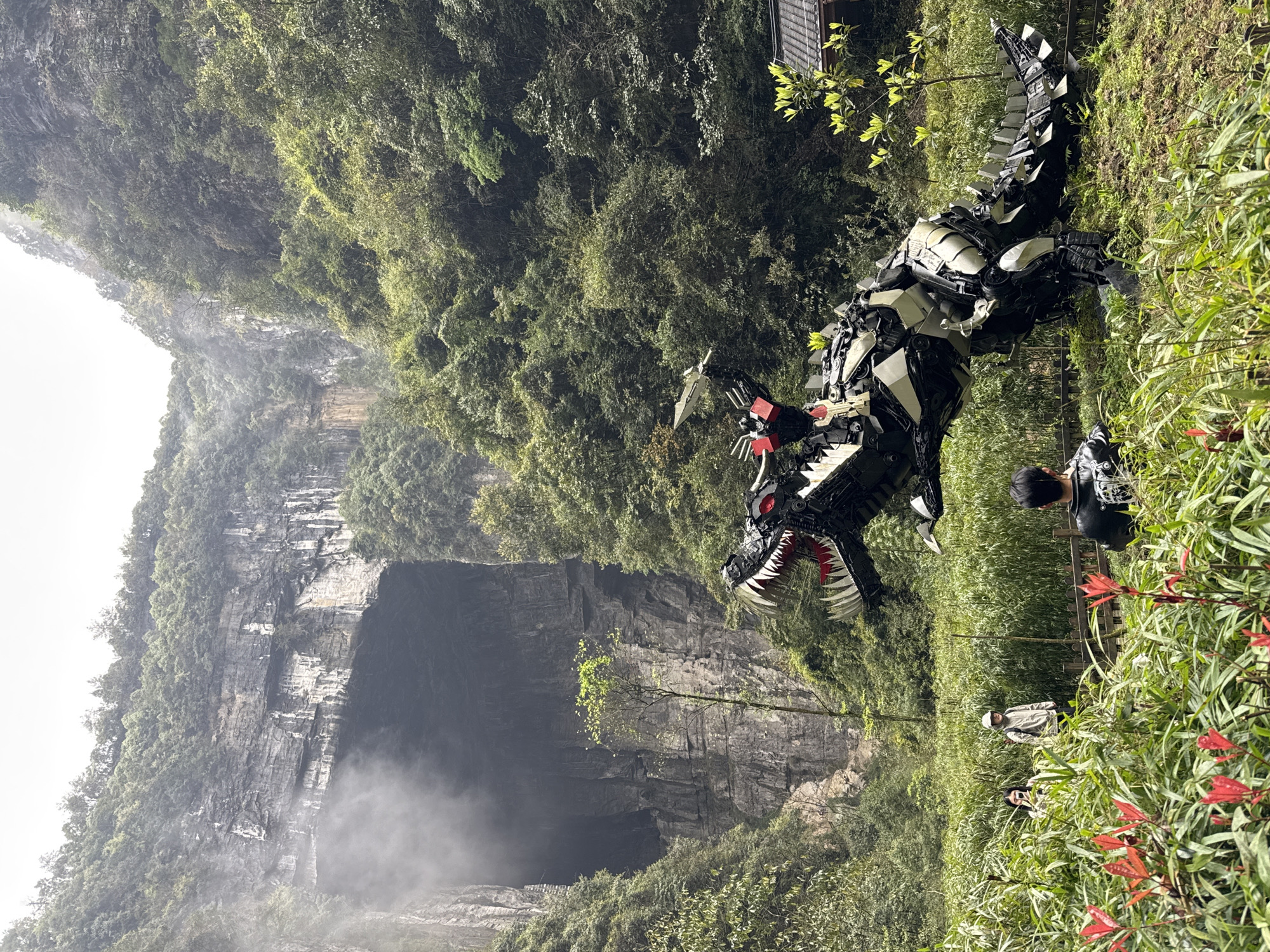 The image depicts a large, metallic dinosaur statue positioned near a deep sinkhole in the city of Wulong District, China. The statue is primarily black and silver with red accents around the eyes and mouth. It is positioned amid dense green vegetation, with bamboo and other plants visible. To the right of the dinosaur, there are several people standing along a wooden fence. They appear to be tourists, observing the statue and possibly the surrounding landscape. In the background, a towering rock cliff forms one side of the sinkhole, which is partially obscured by mist or fog. The overall environment is lush and mountainous.