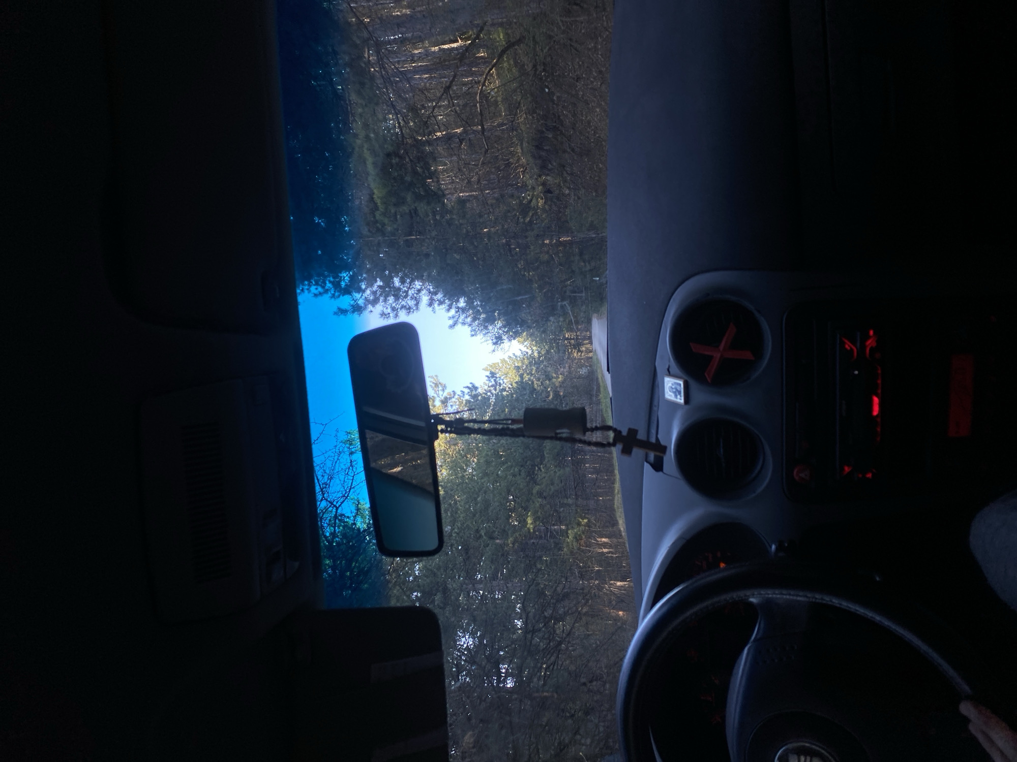 The image shows the interior of a car, specifically the driver's seat perspective, looking out through the windshield. The car is on a road surrounded by trees. A rearview mirror is visible in the center, with a small crucifix hanging from it. The dashboard is also visible, including the steering wheel, radio, air vents, and other controls. The location is unknown; no information is available to place this scene in Blagoevgrad, Bulgaria.