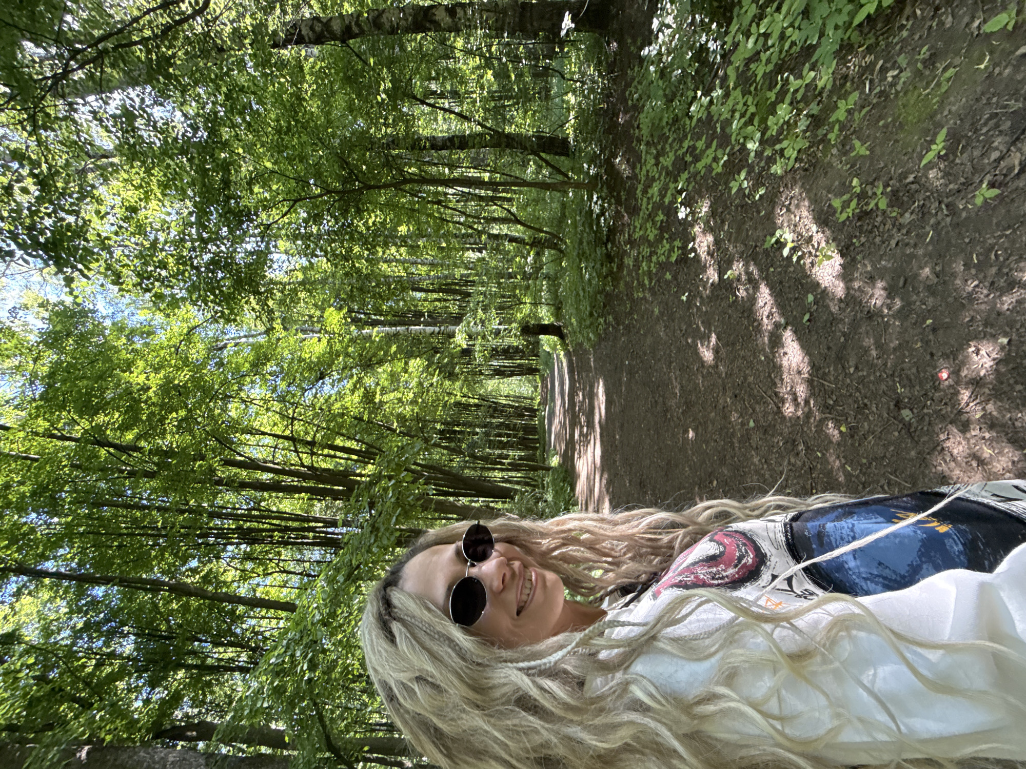A photograph depicts a female subject situated on an unpaved dirt path within a densely forested area. The subject, positioned in the left foreground, is a female with long, blonde, textured hair styled into dreadlocks or twists, and fair skin. She wears dark, round sunglasses and a white long-sleeved top, partially revealing a colorful garment with an abstract blue, black, white, red, and orange pattern. Her head is turned towards the camera, and she exhibits a clear smile. The path recedes into the background, flanked by numerous deciduous trees with full green foliage, indicative of a spring or summer setting. Sunlight filters through the tree canopy, creating dappled light and shadows on the path and ground vegetation, which consists of small green plants and leaf litter. The scene is located in Rusanovo, Russia, presenting a bright, daytime outdoor environment.