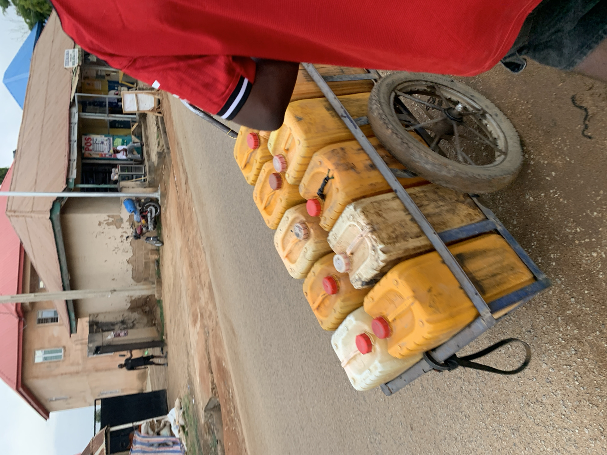The image shows a metal cart filled with yellow and white plastic jerrycans, many with red caps. The cart has one visible spoked wheel. A person wearing a red shirt with white and black trim is partially visible, pulling the cart on a paved road in New Stadium, Nigeria. To the left of the road, there are single-story and two-story buildings with brown roofs. A person dressed in black is visible near one of the buildings. A motorcycle is parked near the entrance of a building further down the road. The sky is overcast.