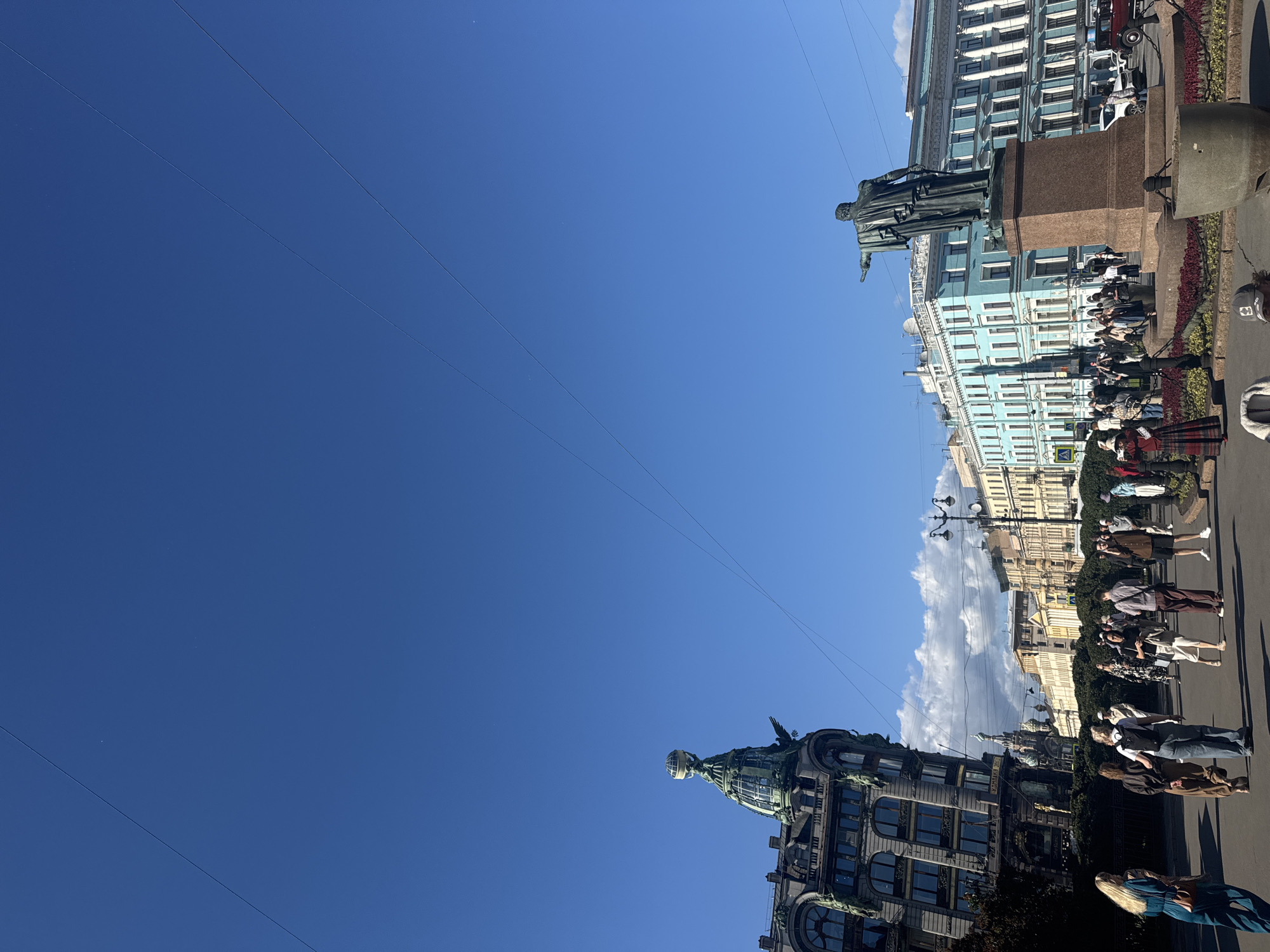 This media file captures a street scene in Saint Petersburg, Russia, under a clear blue sky. The image is oriented such that the buildings and sky are on the right side, and the majority of the frame is filled with the vast expanse of the sky, with several thin wires crisscrossing it.
On the right side of the image, a statue of a man stands on a pedestal in front of a light blue building with classical architecture. A crowd of people is gathered on the sidewalk in front of the building, appearing to be observing something or engaging in a guided tour. They are casually dressed, suggesting a warm day. Further down the street, more buildings with ornate facades are visible, as well as streetlights and greenery.
On the left side, a darker, more imposing building with a domed structure at its peak is seen. This building also features intricate architectural details. A few people are visible near this building as well, some standing, others seated or reclining.
The overall impression is of a bright, sunny day, likely daytime given the clear sky and the activity on the streets. The presence of a statue and grand architecture suggests a significant historical or cultural location. The mood appears relaxed and observational, with people taking in the sights. No discernible text is visible in the image.