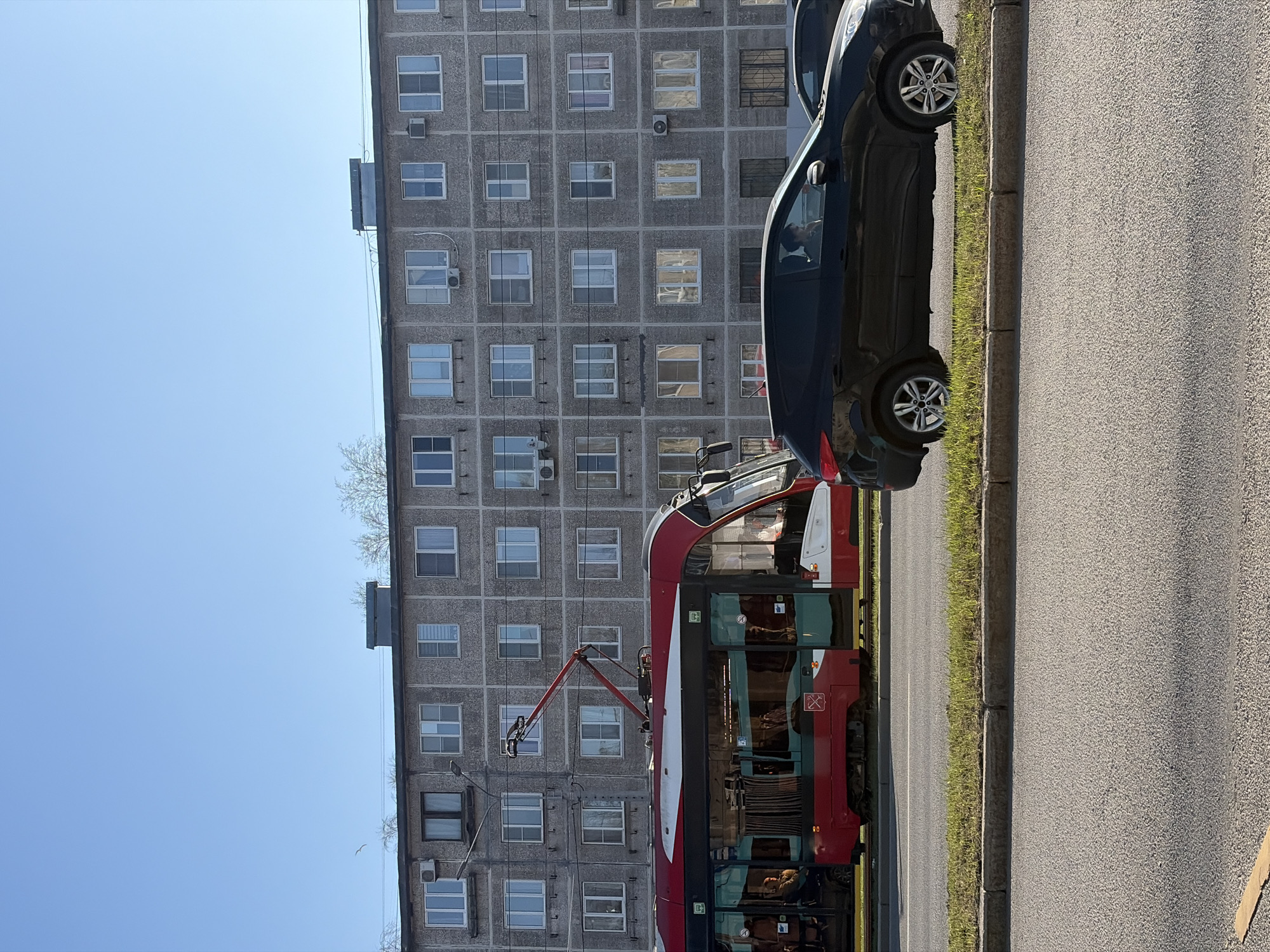 A street scene in Saint Petersburg, Russia, shows a drab, gray apartment building as a backdrop. In the foreground, parallel to the building and along the edge of a paved road, are a black car and a red and white trolleybus. The sky above the building is clear and blue. The trolleybus is connected to overhead wires. A narrow strip of green grass separates the vehicles from the road. The image appears to have been taken during daylight.