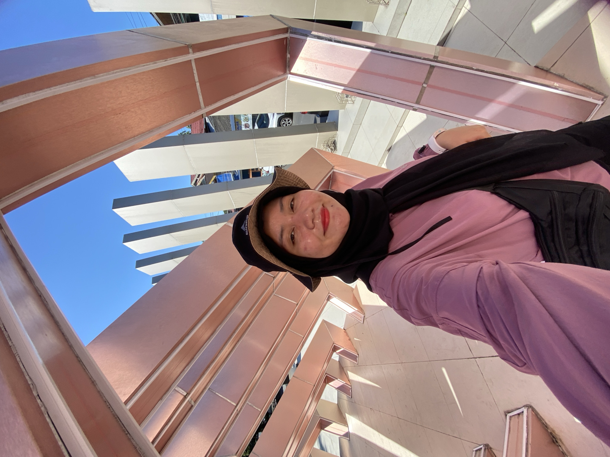 A woman takes a selfie outdoors in front of a modern architectural structure. She is wearing a pink long-sleeved shirt, a black hijab, and a bucket hat. The structure is composed of a series of angled metallic beams, some with a copper or rose-gold finish and others in a light gray or white. The background shows a clear blue sky and some buildings and trees, suggesting an urban or semi-urban environment. The ground is paved with light-colored tiles. Based on the context, this location is likely in Banjarbaru, Indonesia.