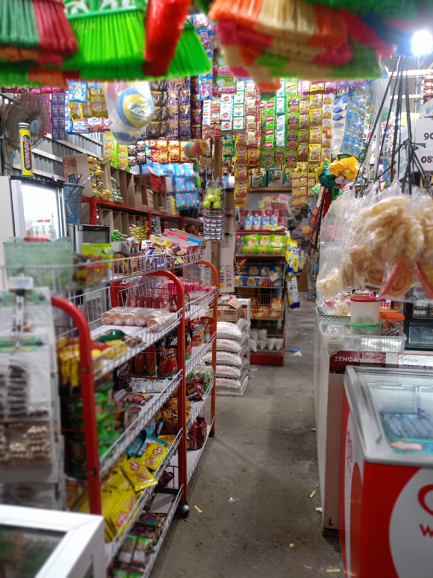 The image shows the interior of a small convenience store, likely located in Uteunreutoh, Indonesia, judging by the branding of items and text visible in the image. The store is packed with various goods, with colorful plastic brooms hanging from the ceiling at the top of the frame. Below the brooms, there's an array of hanging single-serving snack packets. The shelves are stocked with items such as snacks, packaged food, drinks, and household goods. A refrigerator with red and white branding is visible on the right side of the image. Sacks of goods are stacked on the floor in the middle of the aisle. The overall impression is one of a crowded and well-stocked neighborhood store.