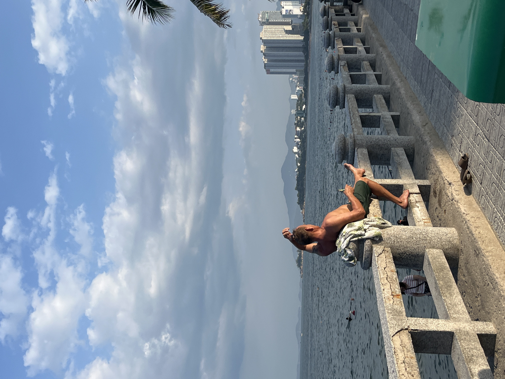 This image depicts a coastal scene in what is identified as Bac Nha Trang Ward, Vietnam. In the foreground, a Caucasian man, shirtless and wearing olive green shorts, sits casually on a weathered concrete seawall railing with his bare feet resting on the top bar. He is tanned, has short light-colored hair, and a visible tattoo on his left bicep. His right hand is positioned at the back of his head, while his left hand holds a green bottle. A white and green patterned shirt is draped over the railing next to him. A pair of dark flip-flops rests on the tiled promenade directly beneath his feet.
The concrete railing, showing signs of wear and cracking, extends horizontally across the mid-ground, separating the promenade from the sea. Through the gaps in the railing, a person wearing a striped shirt is partially visible, facing the water. The sea, a deep blue-green, occupies the middle distance, with several indistinct figures swimming or wading. One person in the far left of the water appears to be wearing a red cap. A small white boat is visible on the distant water to the left.
Beyond the water, a cityscape with numerous modern high-rise buildings lines the coast, backed by a range of hazy mountains under a partly cloudy sky. The sky transitions from a lighter blue with scattered white cumulus clouds at the top to a denser band of gray clouds near the horizon. The fronds of a palm tree are visible in the upper right corner. A large, dark green, possibly curved object is present in the lower right foreground on the tiled promenade. The lighting suggests late afternoon or early morning, with a warm glow on the man.