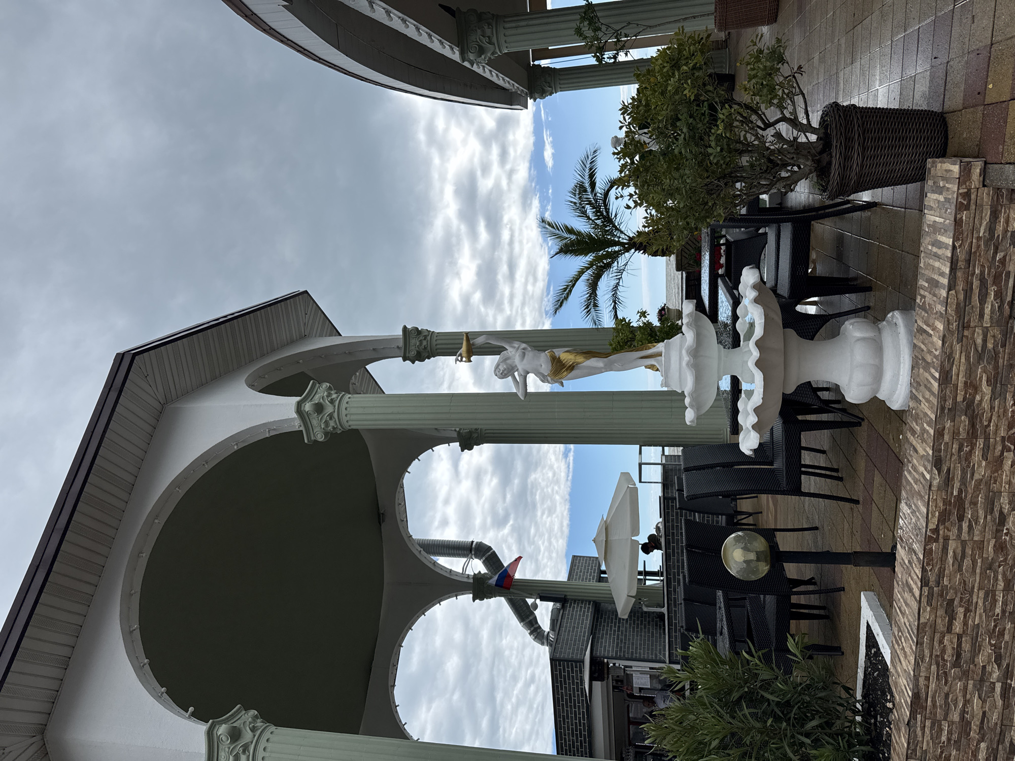 The image shows an outdoor area in Sochi, Russia. It features an open structure with white columns and arches. A white statue of a figure holding an object is visible, along with a decorative white fountain. Tables and chairs are arranged nearby. A palm tree is partially visible in the background. The sky is overcast with clouds. A Russian flag is also seen.