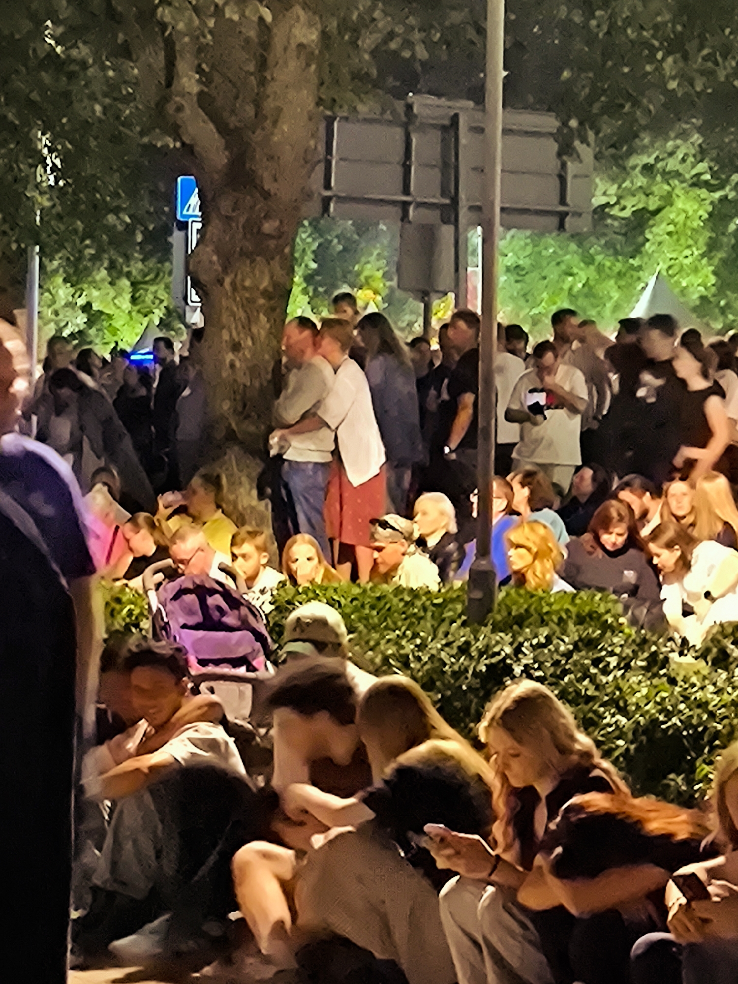 The image shows a crowd of people gathered outdoors at night, with a large tree in the center. The atmosphere appears to be social, with some individuals in close proximity to each other. Many people are looking at their phones. The presence of streetlights and illuminated foliage suggests an urban environment. The image does not contain enough visual information to determine the specific location in Riga, Latvia.